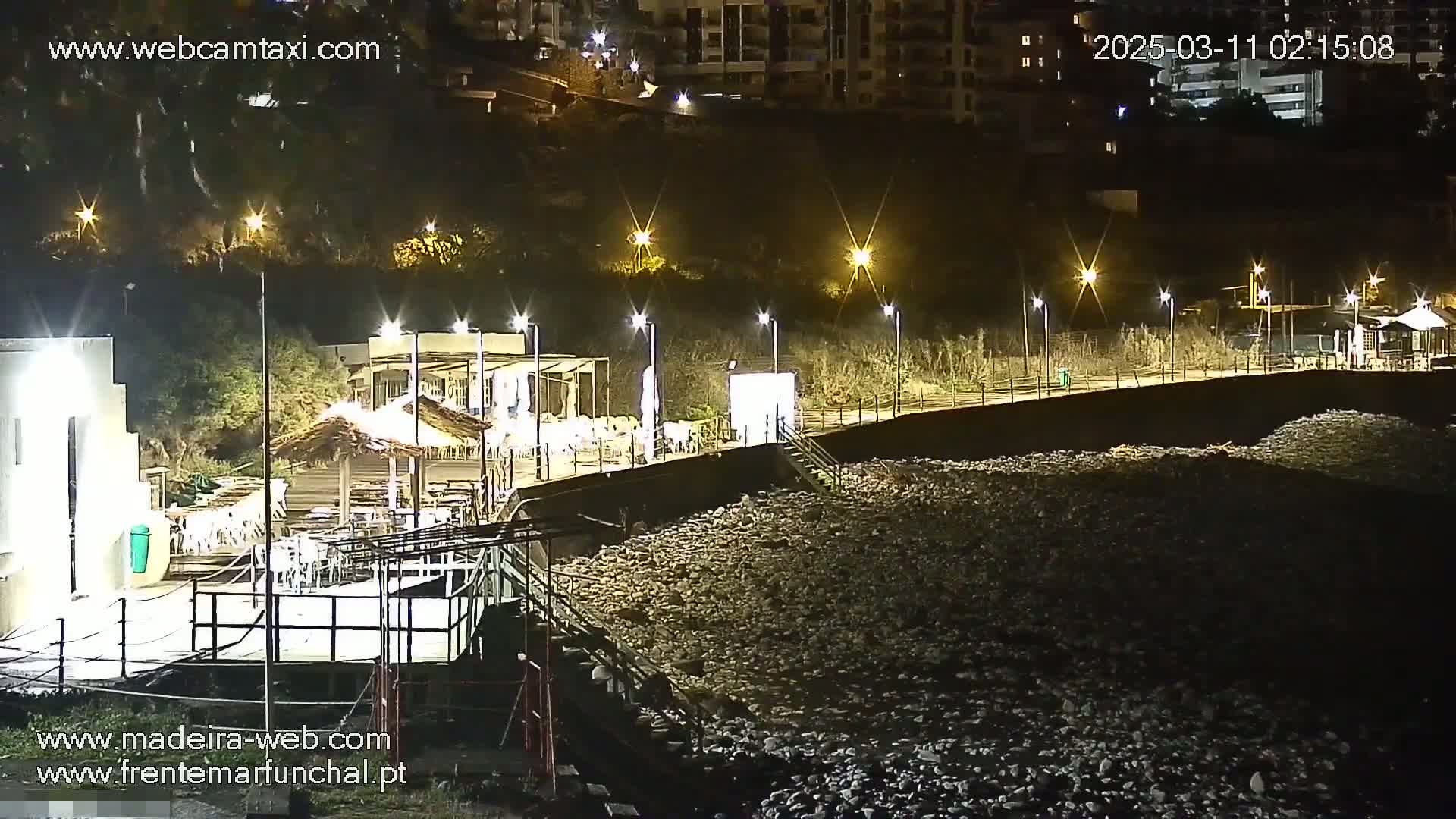 A nighttime view of a rocky beach with illuminated outdoor seating areas and buildings along a seawall, under clear skies.