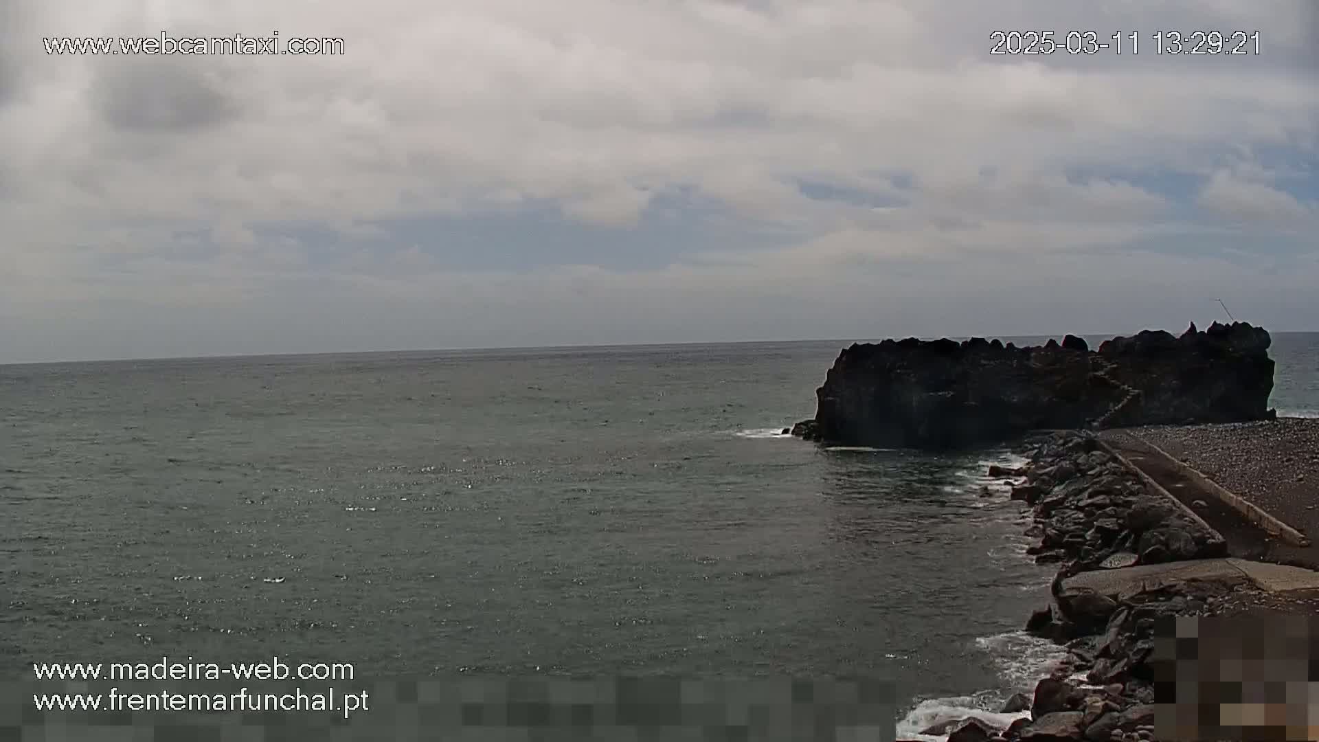 A dark rocky outcrop juts into a calm, gray sea under a mostly cloudy sky.