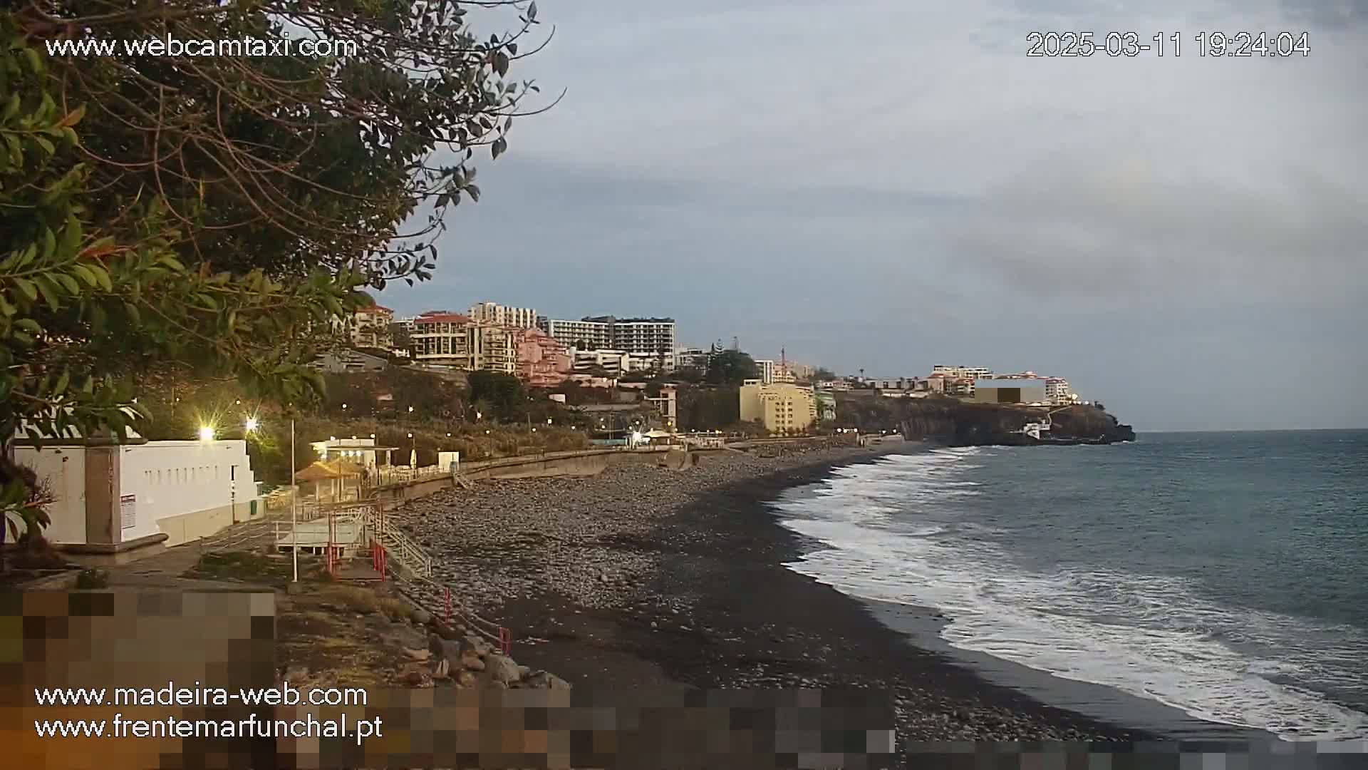 A dark, pebble beach with gentle waves under an overcast sky, backed by a row of buildings and vegetation.