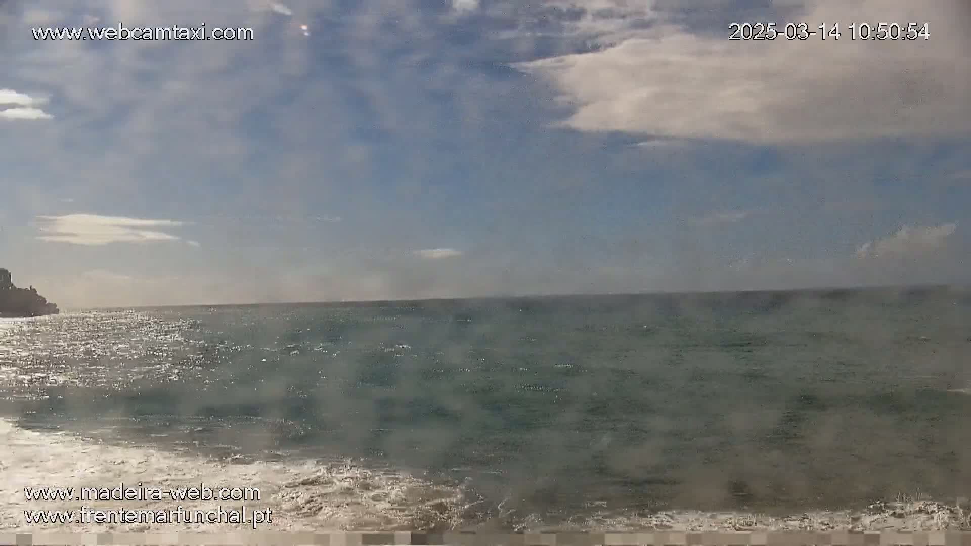 A partly cloudy day shows a view of a dark-green ocean meeting a sandy shore, with a dark cliff visible in the distance.