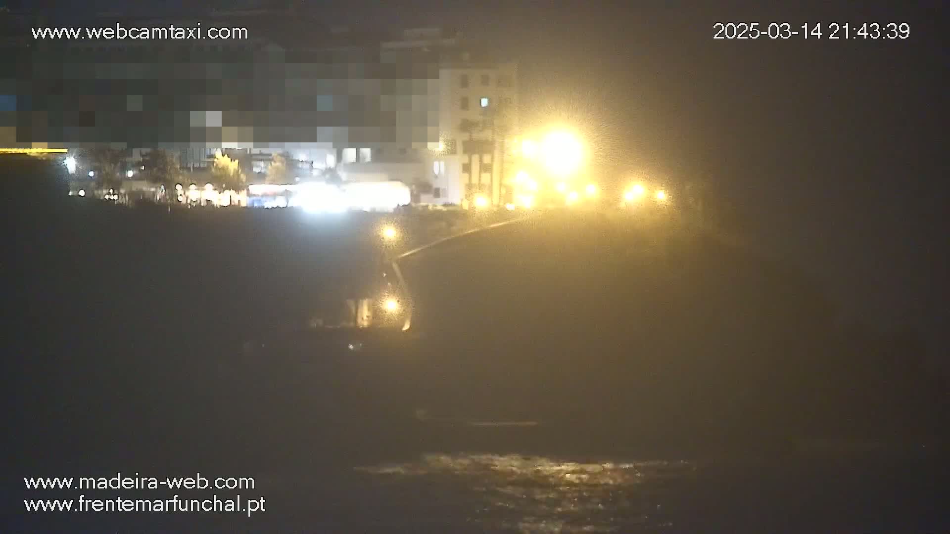 A nighttime, foggy view of a coastal road with buildings and streetlights, overlooking a dark body of water.
