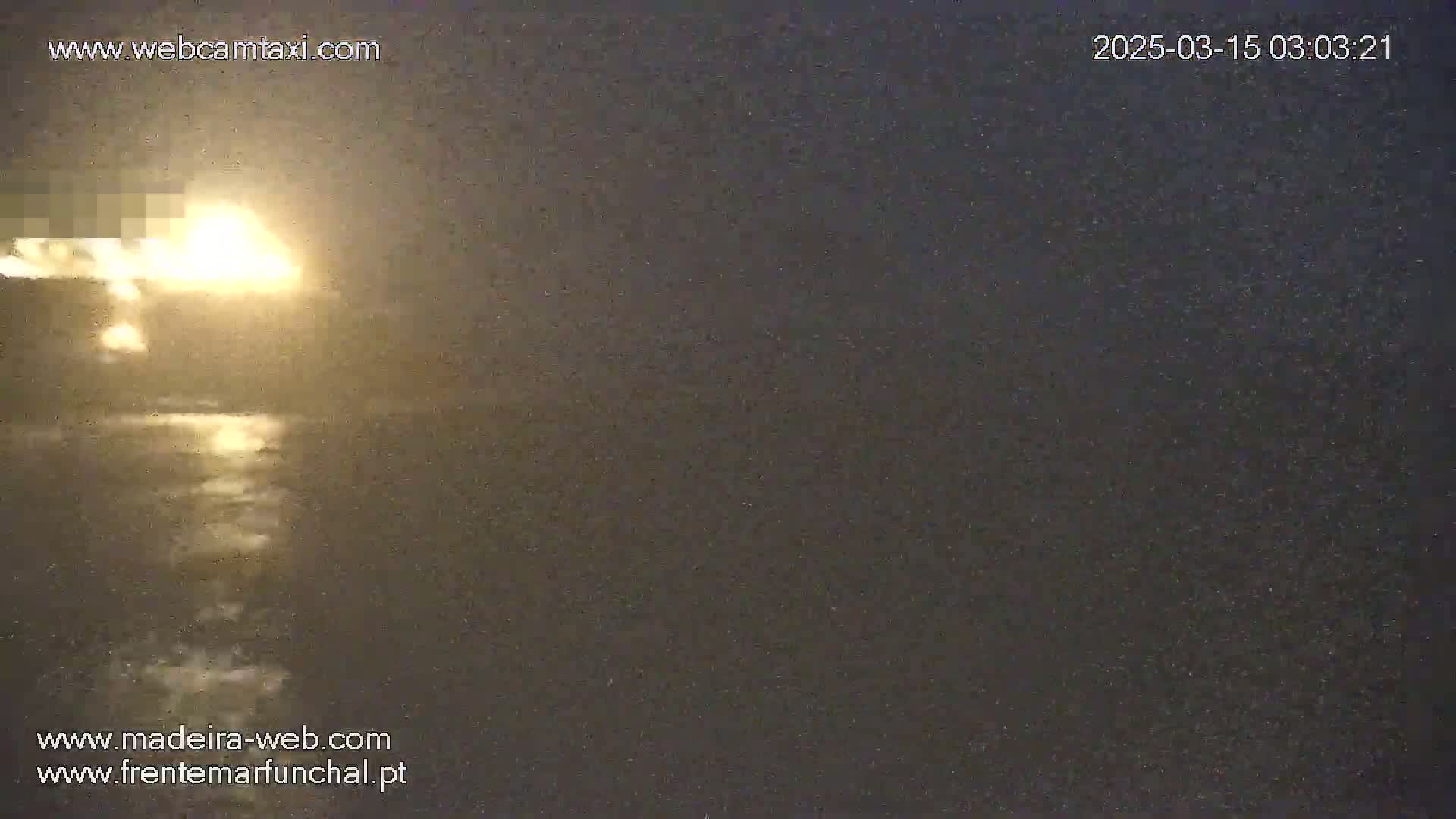 A bright light source, possibly a fire or explosion, is visible on the horizon over a dark body of water under a nighttime sky with light precipitation.