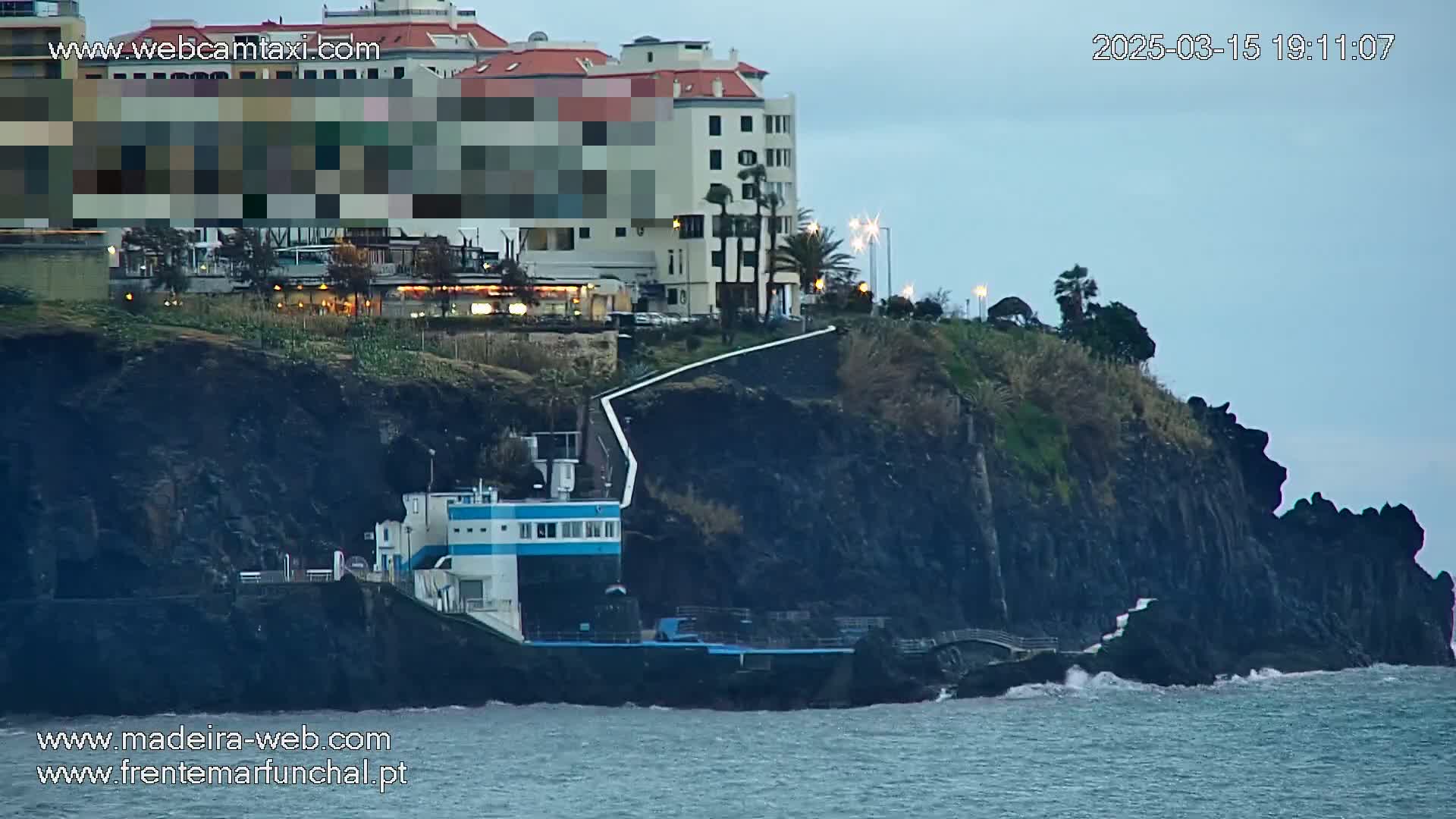 A multi-story building sits atop a dark cliff overlooking a body of water with small waves under an overcast sky.