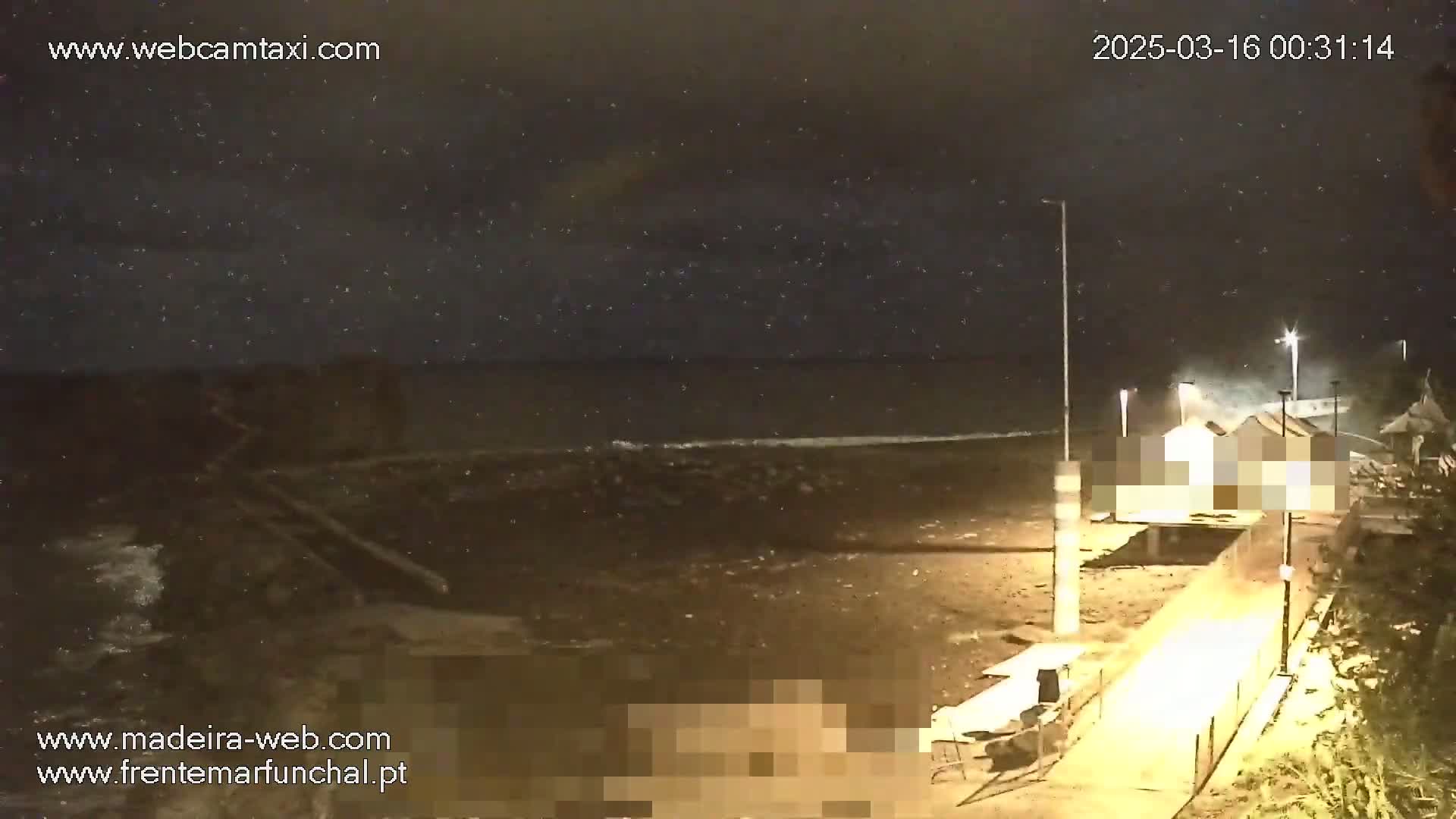 A nighttime seaside scene shows a dark ocean with small waves lapping a sandy shore, a building with obscured portions, and a walkway under a slightly cloudy sky.