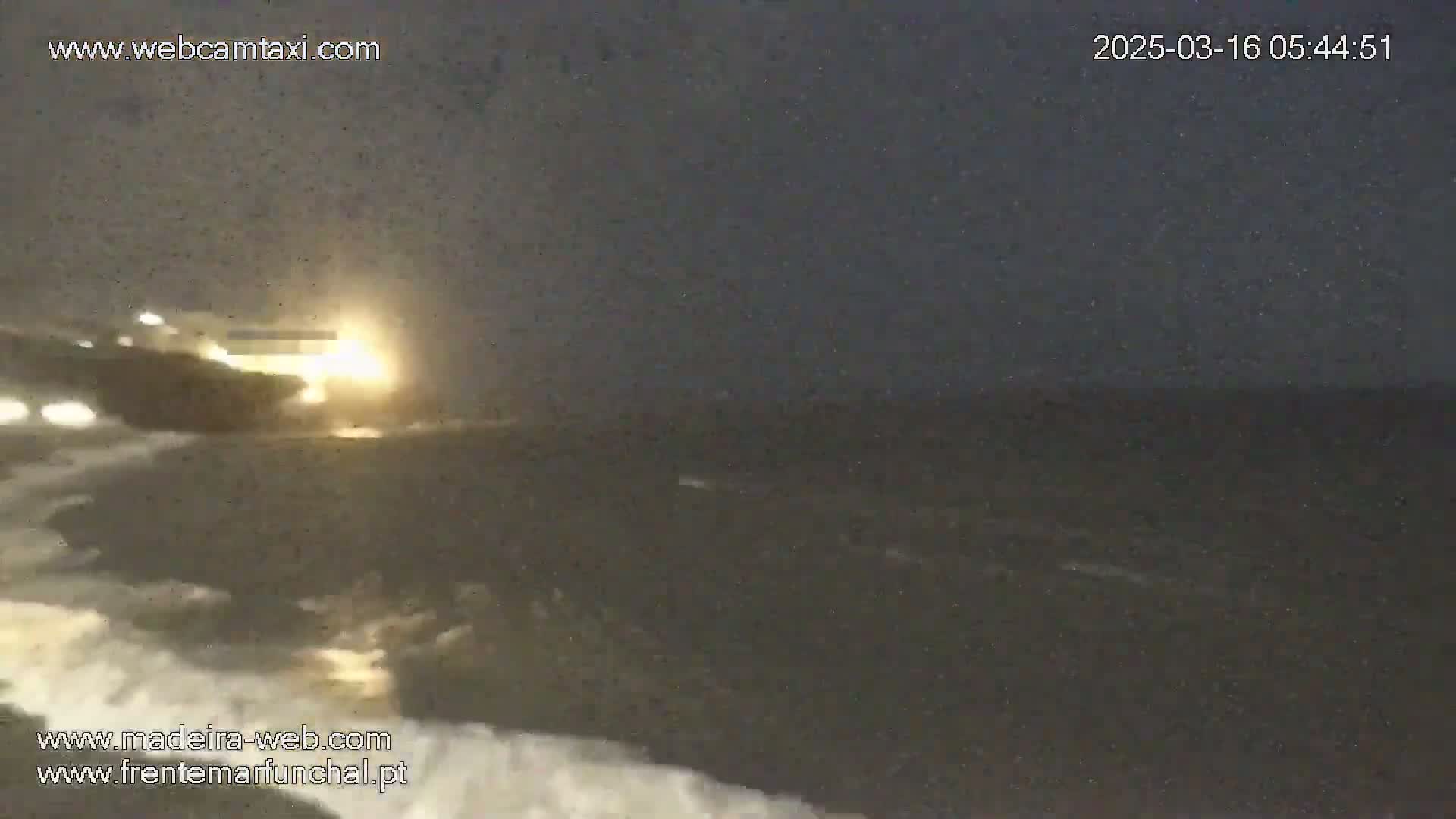 A brightly lit vessel is seen at night on a dark, rough sea with waves breaking against the shore.