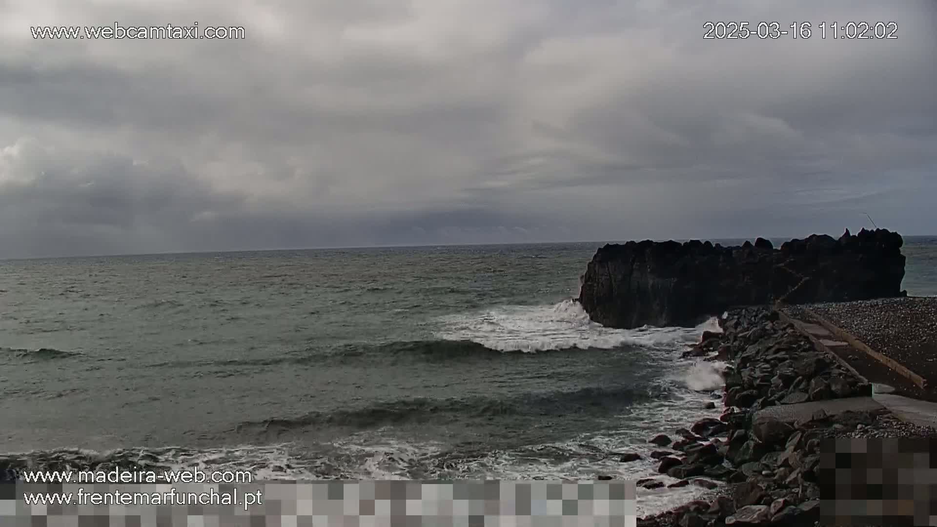 A rocky outcrop in the ocean is partially surrounded by a wave-washed, rocky shore under a cloudy sky.