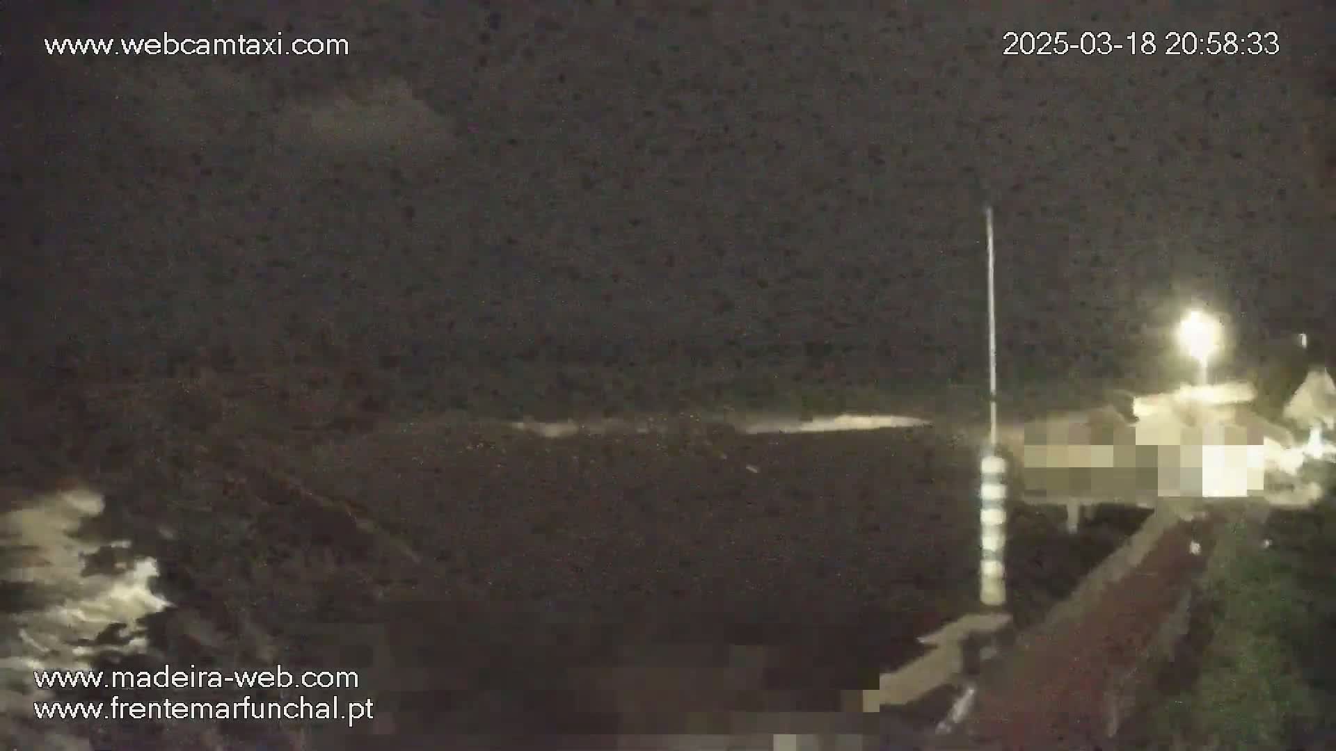 A nighttime, dark ocean scene with some small waves crashing on the shore near a lit-up area, possibly a town.