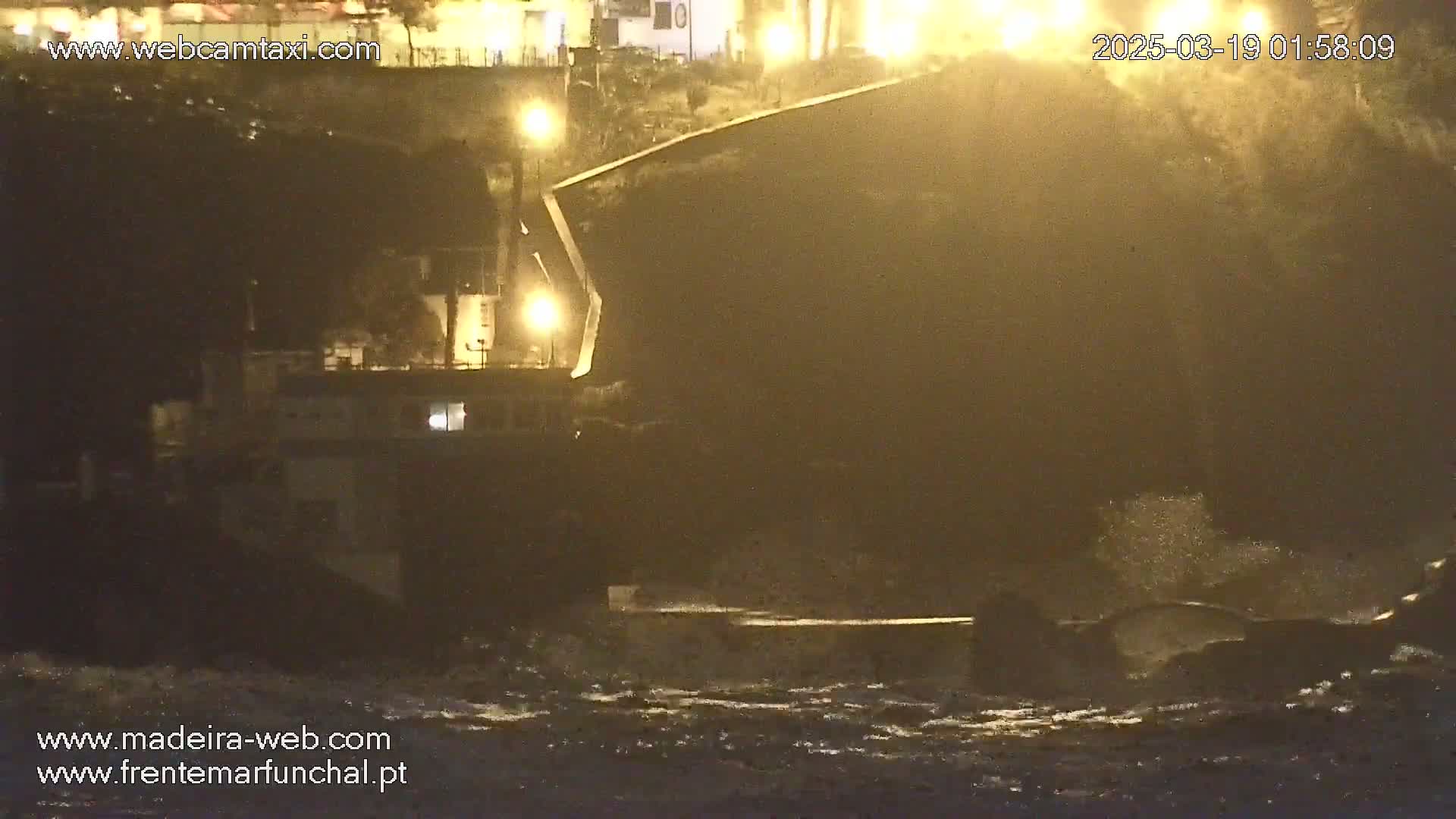 A large dark vessel is moored at a dock at night during a stormy sea with heavy waves.