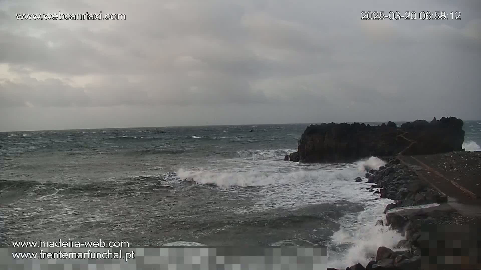 Under an overcast sky, choppy waves crash against a rocky shoreline and a dark, low-lying outcropping of rocks.