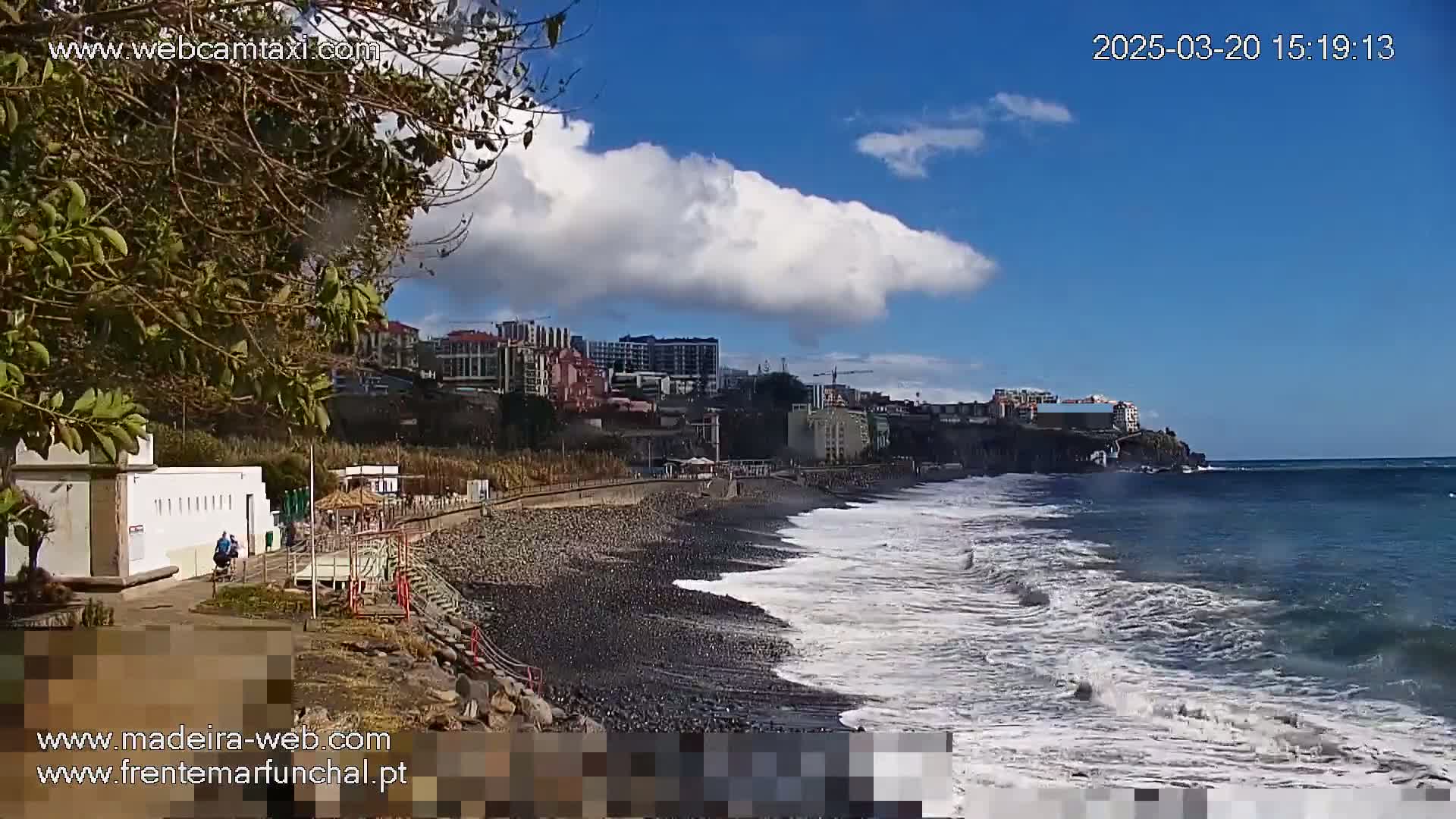 A partly cloudy day shows a dark-sand beach with moderate waves lapping the shore, in front of a city and ocean.