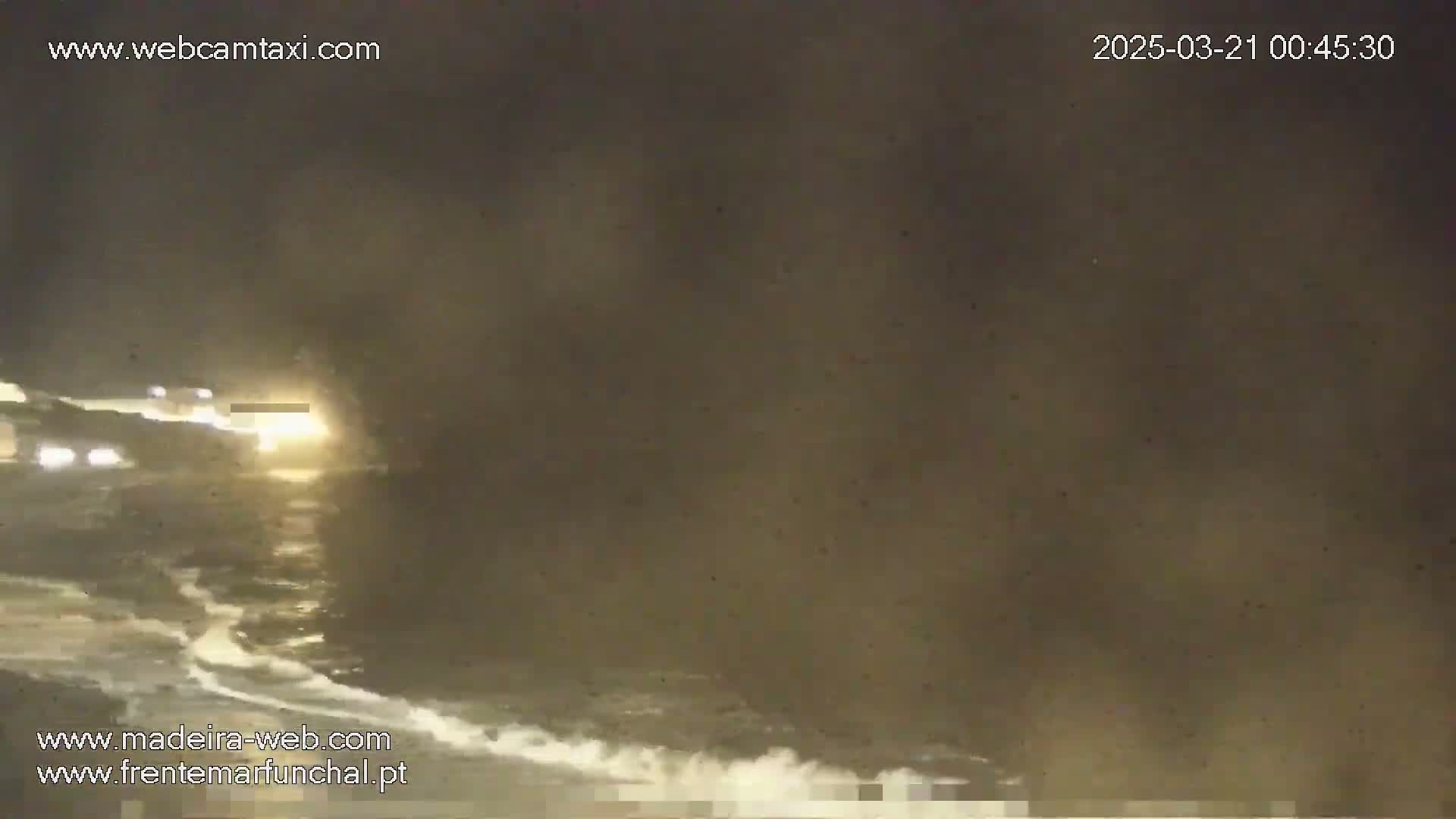 At night, under a hazy, dark sky, waves crash on a shore near a road where vehicles are driving.