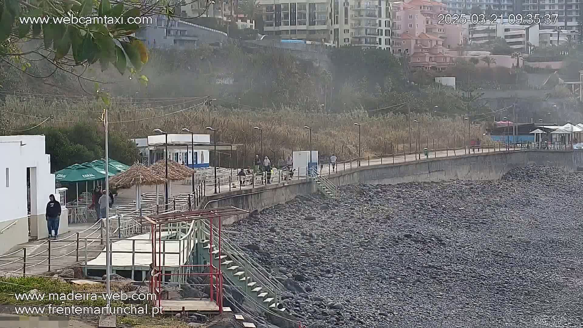 A hazy, outdoor scene shows a rocky beach with a paved walkway lined with small buildings and people, near a hillside with buildings in the background.