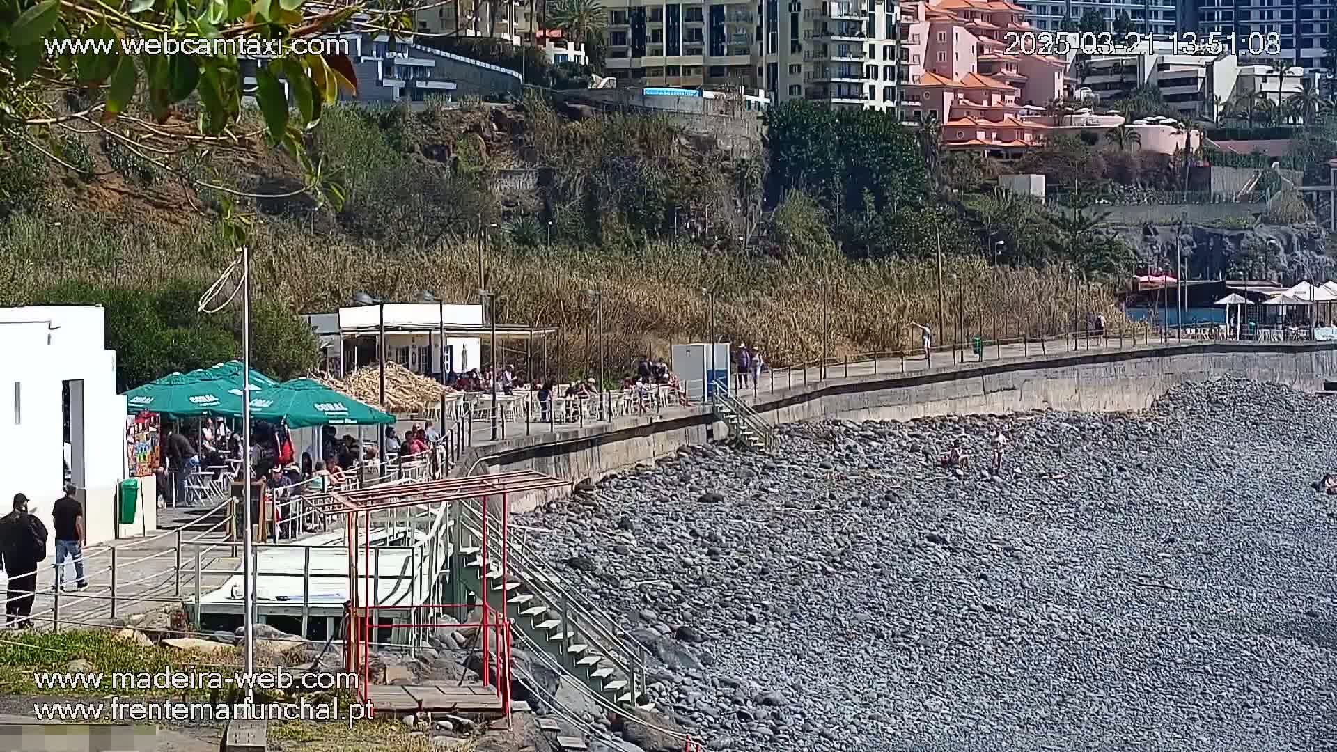 A rocky beach with a paved walkway above it, lined with cafes and people, under a sunny sky.