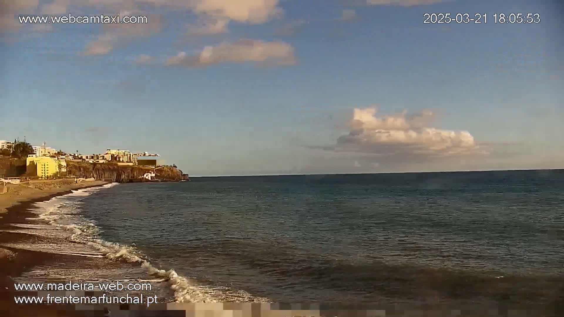 A dark-sand beach with gentle waves laps against a rocky shoreline, under a partly cloudy sky.