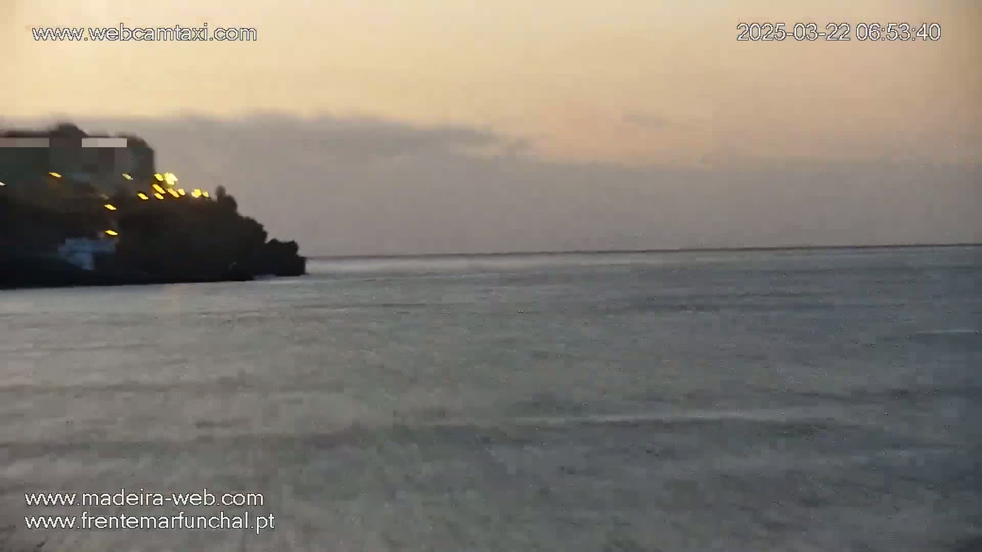 A dark, rocky cliff with lights is visible at the left edge of the image, overlooking a calm, gray-blue ocean under a dusky, overcast sky.
