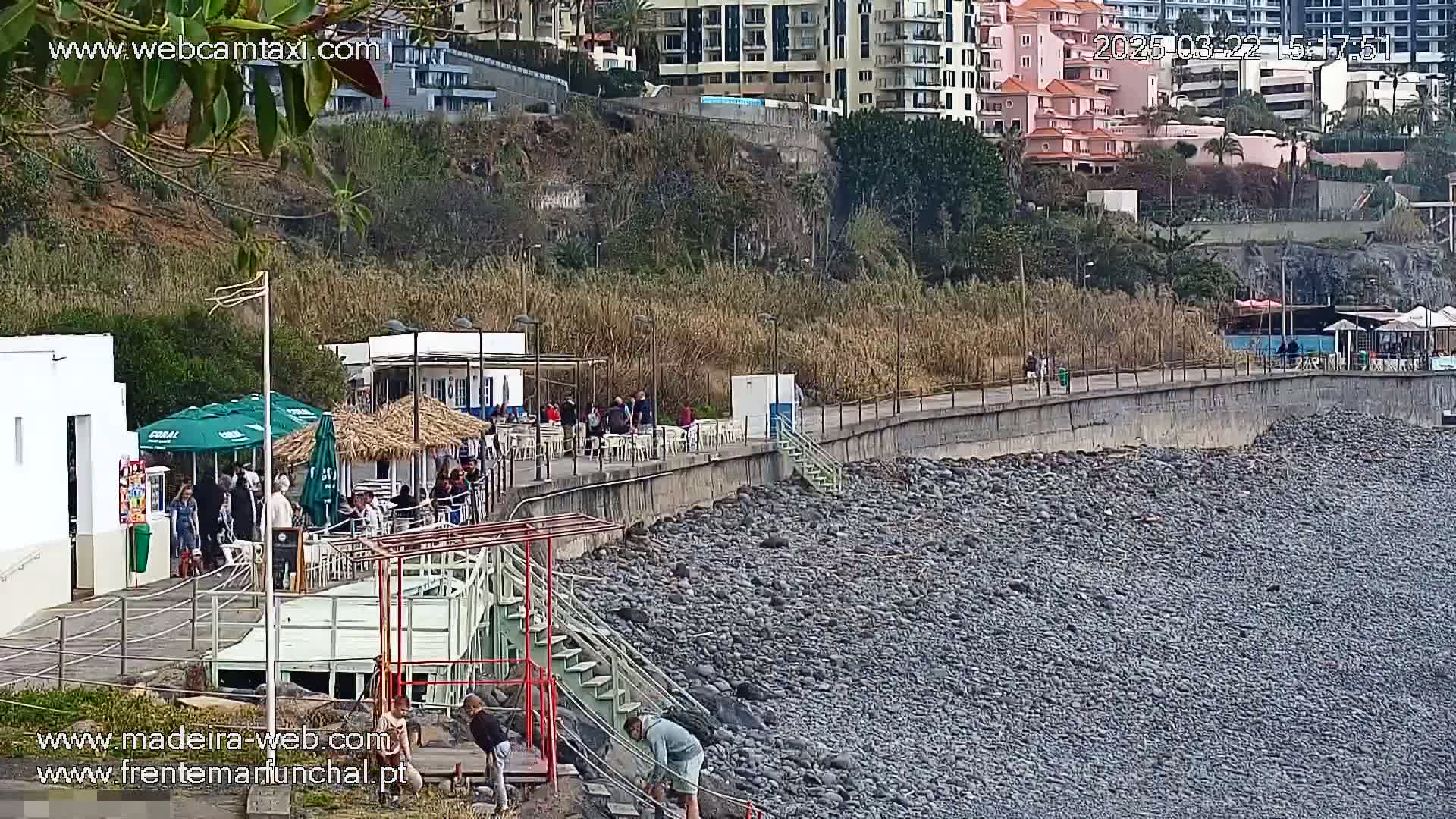 A rocky beach with a paved walkway lined with restaurants and buildings, backed by a hillside with apartments, on a mostly sunny day.
