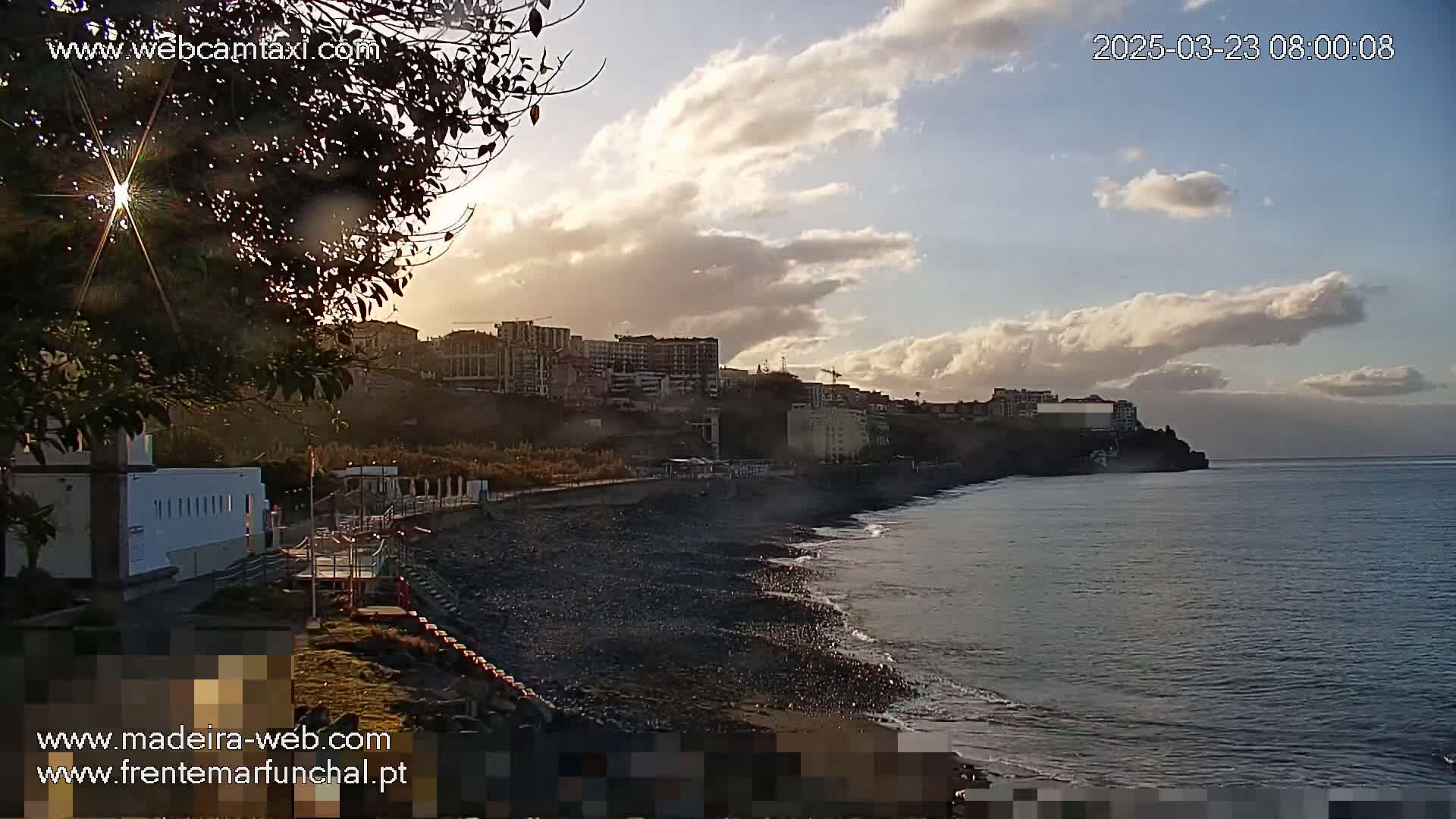 A partly cloudy sunset over a dark-sand beach with buildings on a hill in the background.