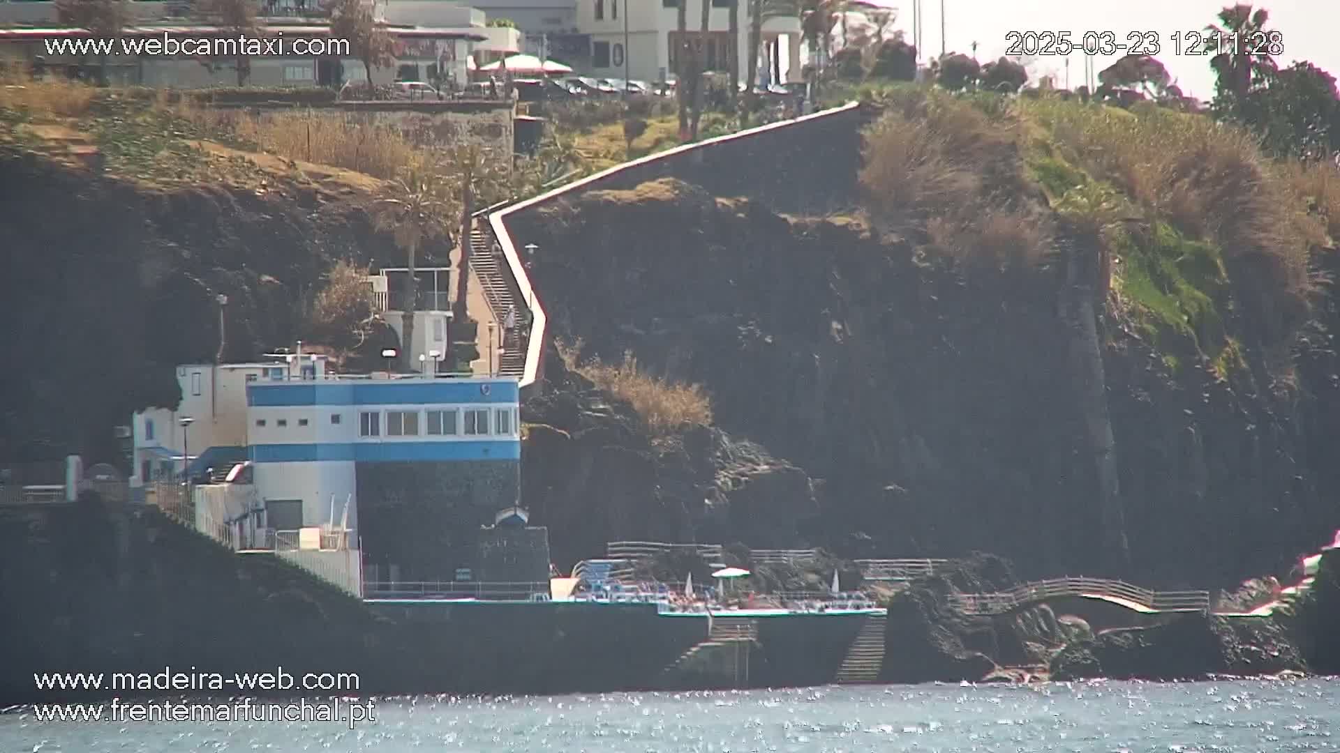 A blue and white building sits near a rocky coastline with a long stairway leading up to a wall, on a sunny day.