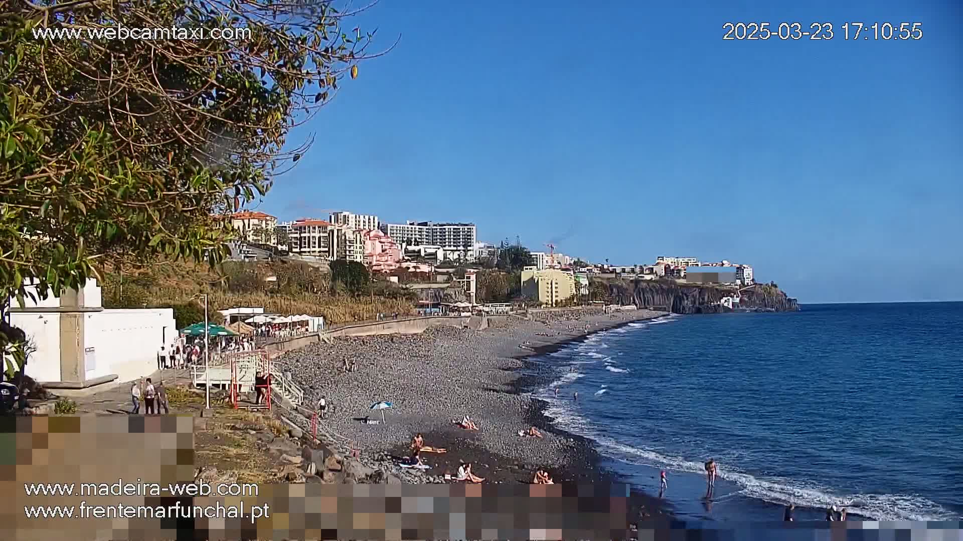 A pebble beach with a few people sunbathing stretches along a coastline under a clear blue sky, backed by buildings and vegetation on a cliff.