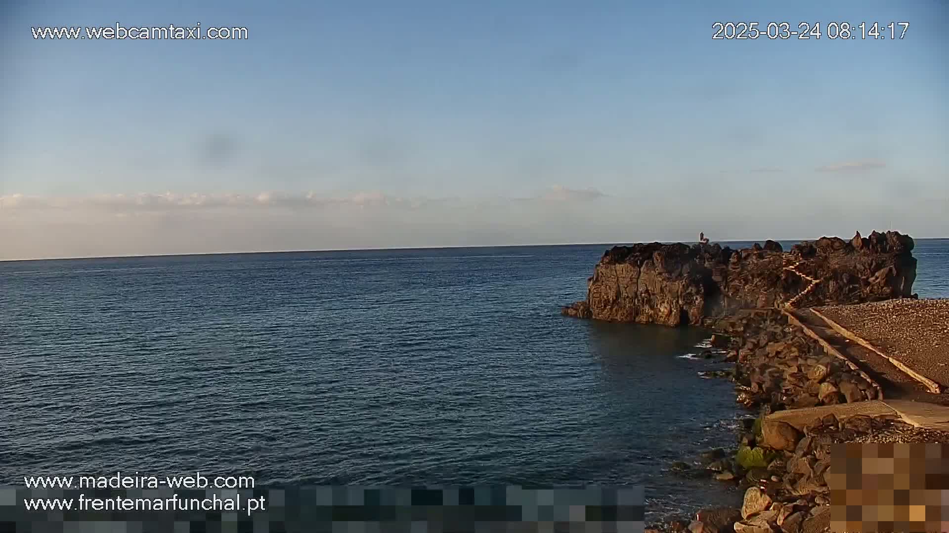 A rocky outcropping with a walkway extends into a calm ocean under a mostly clear sky.