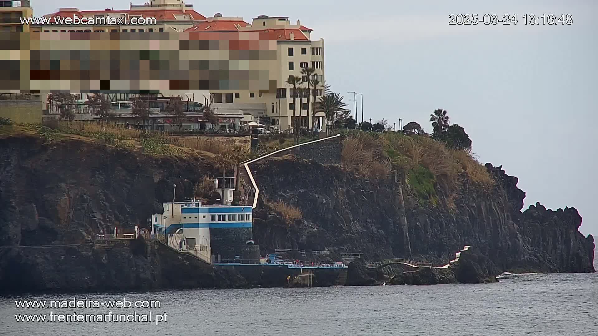 A multi-story building sits atop dark cliffs overlooking a calm ocean, with a walkway and small building along the shoreline on an overcast day.