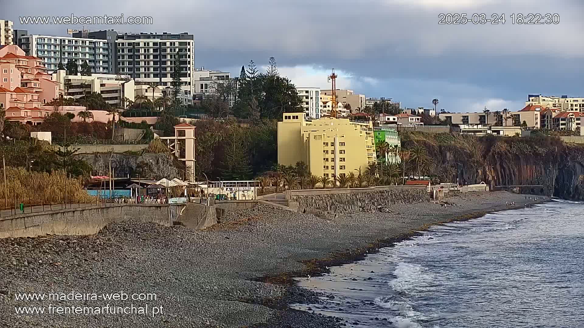 A rocky beach with gentle waves laps against the shore in front of a seaside town under a cloudy sky.