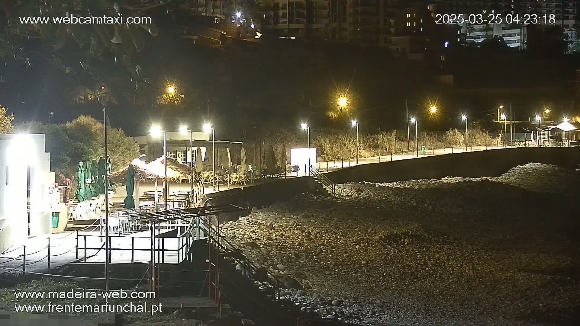 A nighttime view of a rocky beach with illuminated buildings and a boardwalk, under clear skies.