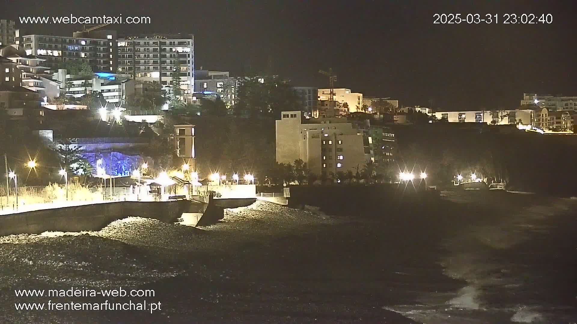 At night, a rocky shoreline with moderately rough waves is illuminated by lights from buildings on a hill overlooking the water.
