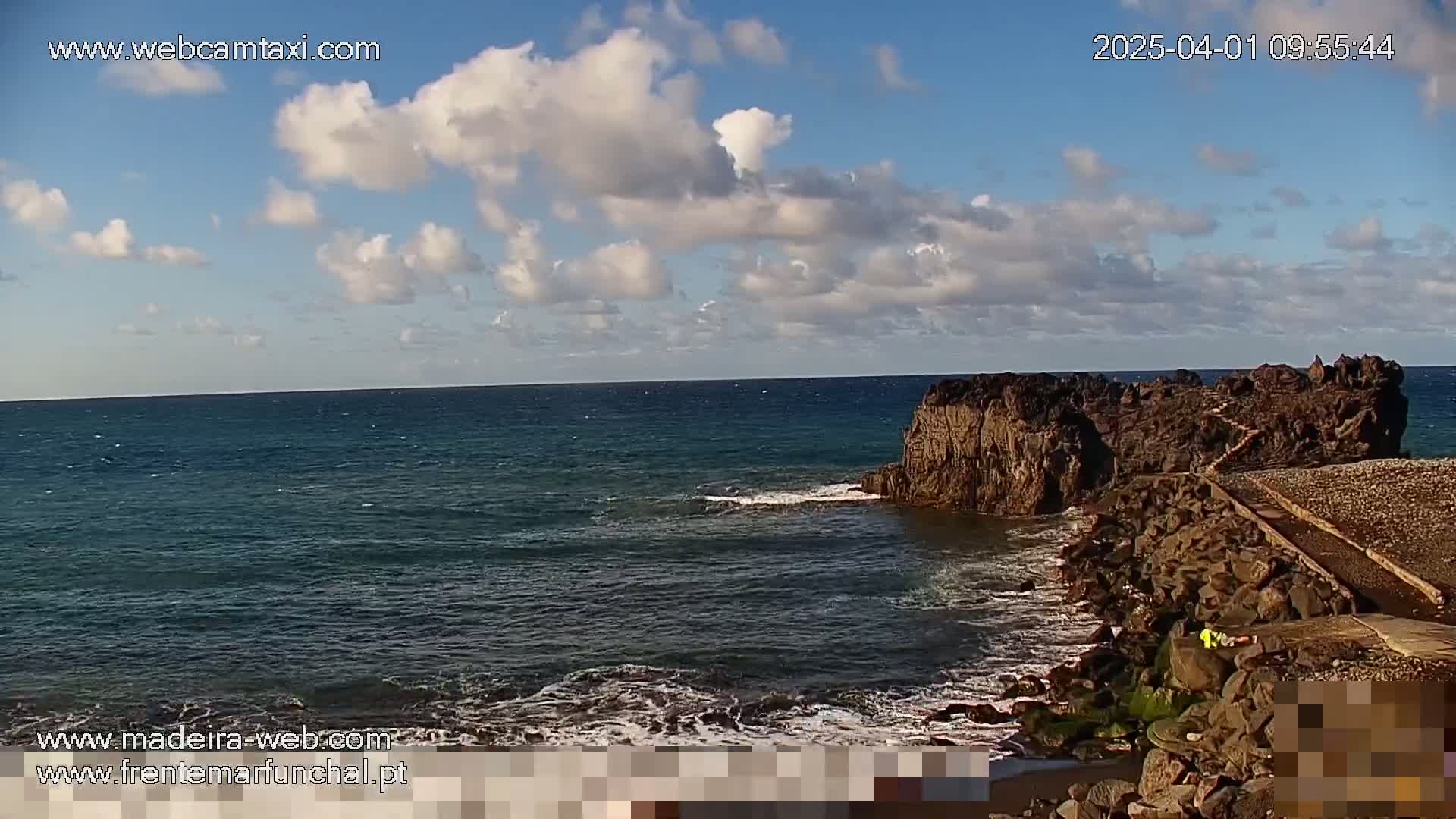 A rocky outcropping juts into a dark blue ocean under a partly cloudy sky.