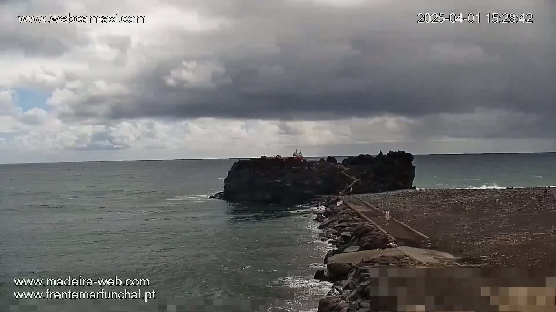 A rocky islet connected to the shore by a narrow path is visible under a cloudy sky, with a dark-grey ocean stretching to the horizon.