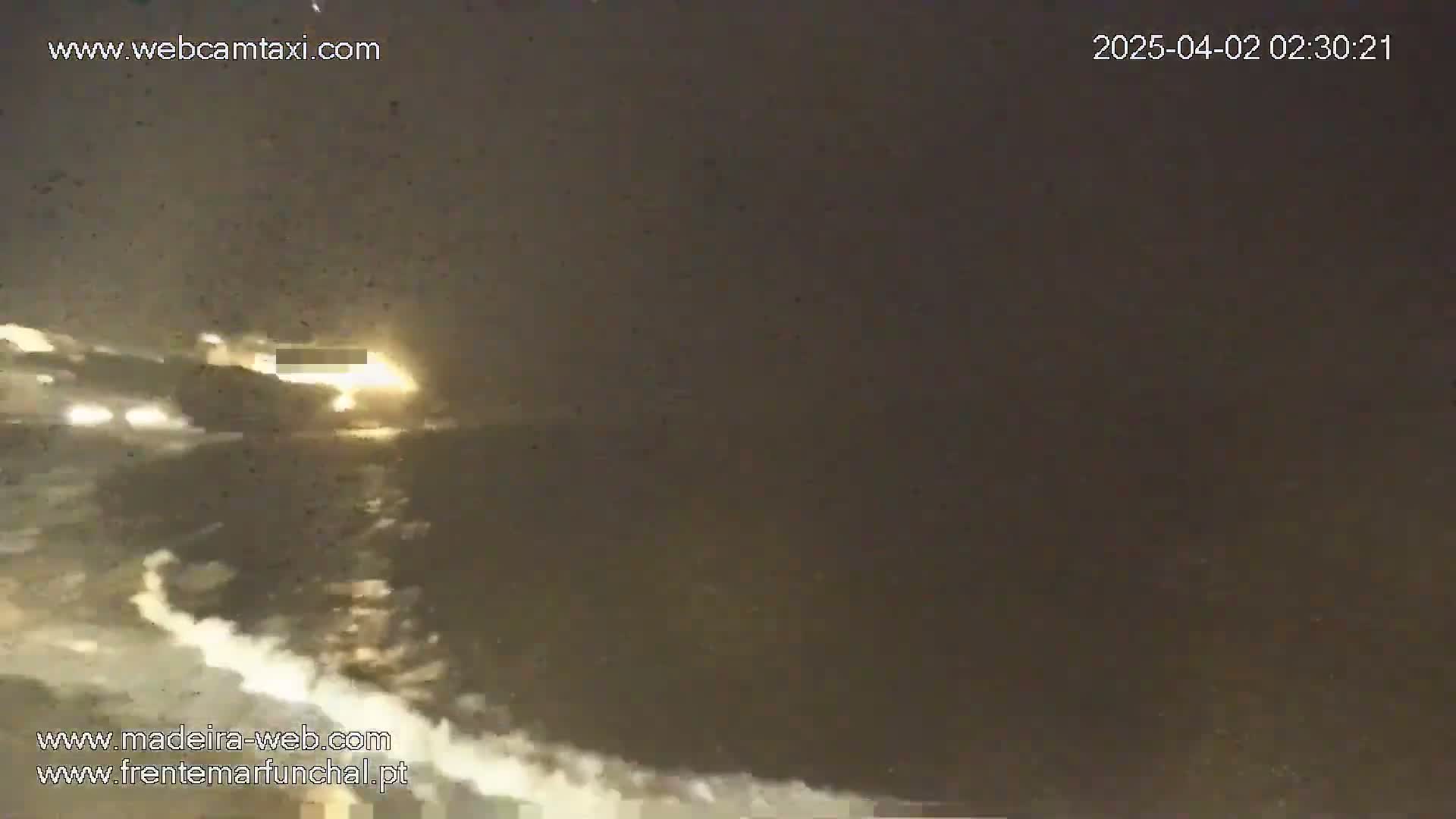 A nighttime, low-light view shows a dark, possibly coastal, scene with vehicle headlights visible in the distance and some white wave-like formations in the foreground.