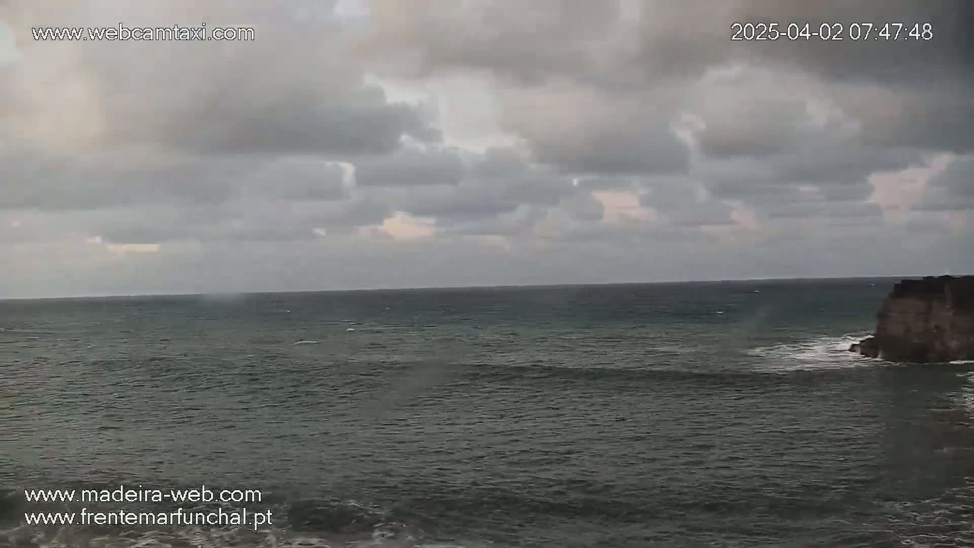 A dark, cloudy sky hangs over a choppy ocean meeting a rocky cliff face.