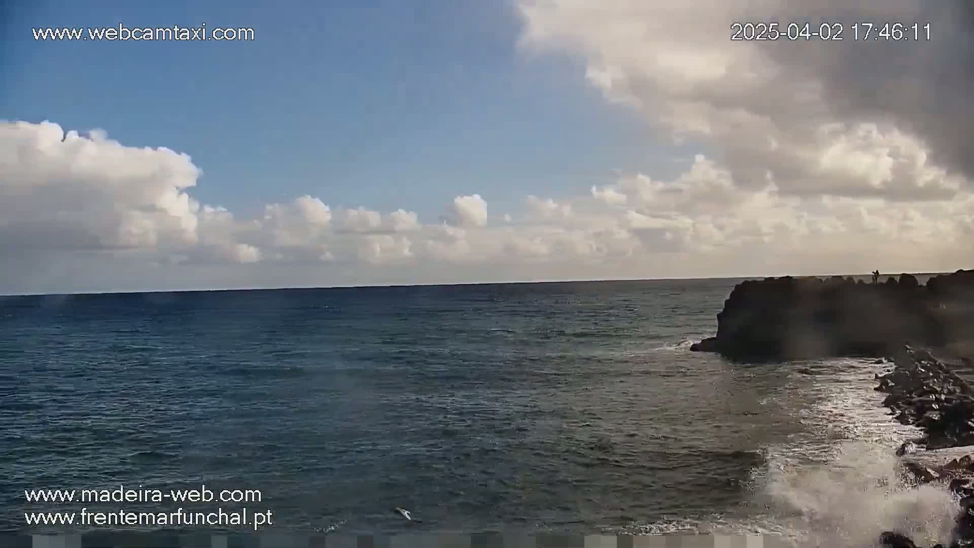 A partly cloudy sky overlooks a dark-blue ocean meeting a rocky shore where small waves crash against the rocks.
