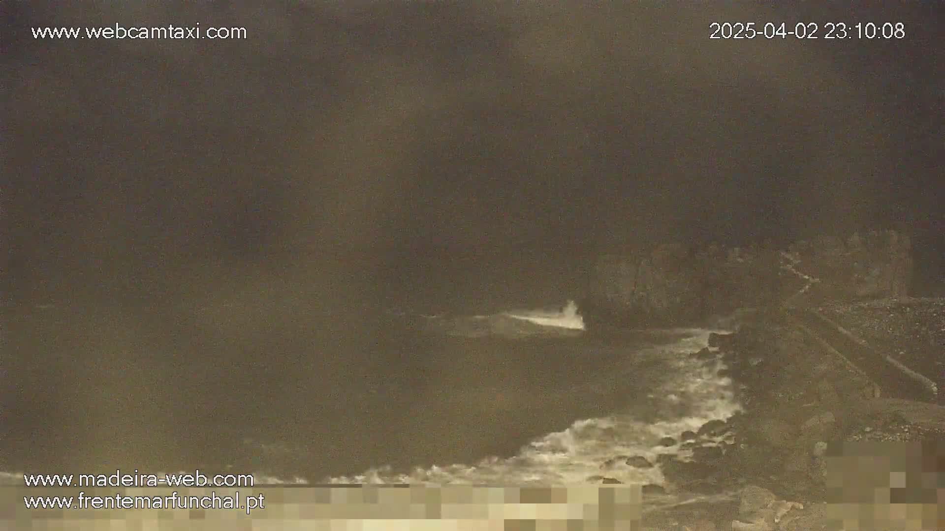 A dark, nighttime coastal scene shows waves crashing against a rocky shoreline under a cloudy sky.