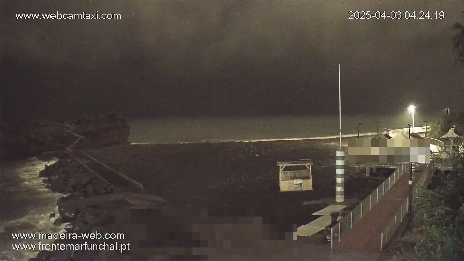 A dark, nighttime beach scene shows a mostly empty black sand beach with a boardwalk, a small structure, and a rocky shoreline with waves crashing against it under an overcast sky.