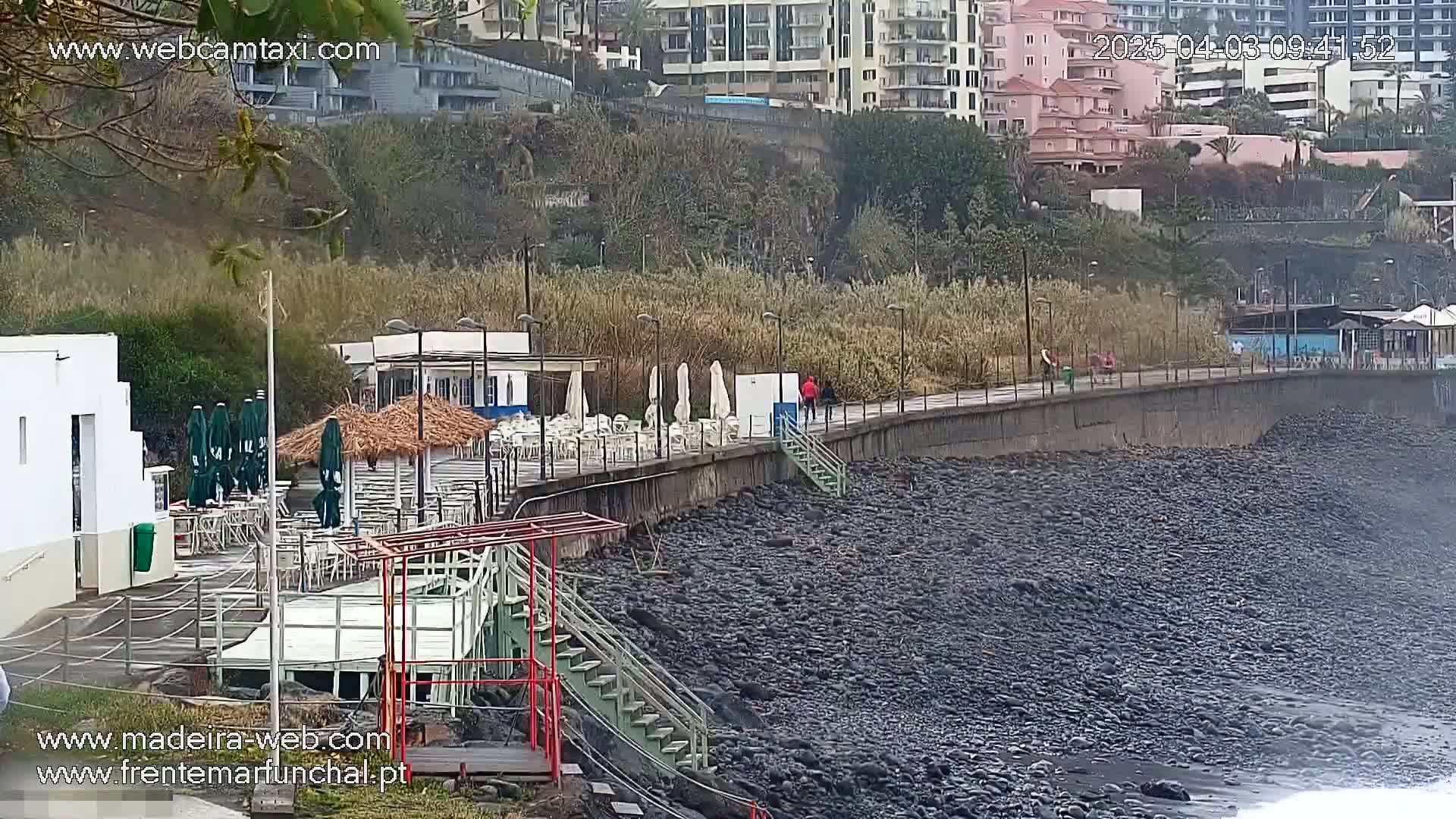 A seaside promenade with outdoor seating areas and a rocky shore is visible on an overcast day.