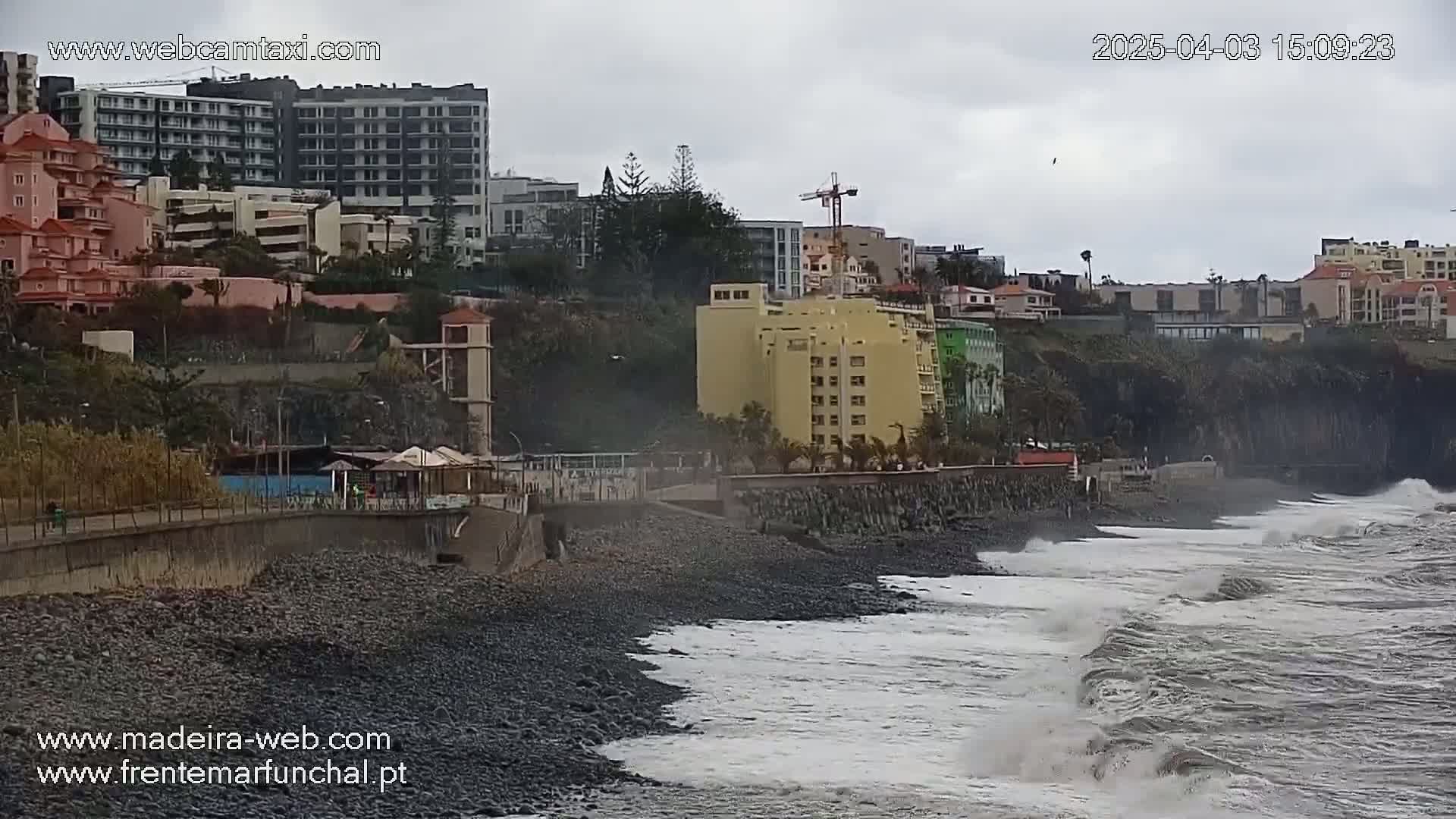 A rocky beach with moderate waves crashes against the shore next to a coastal city under an overcast sky.
