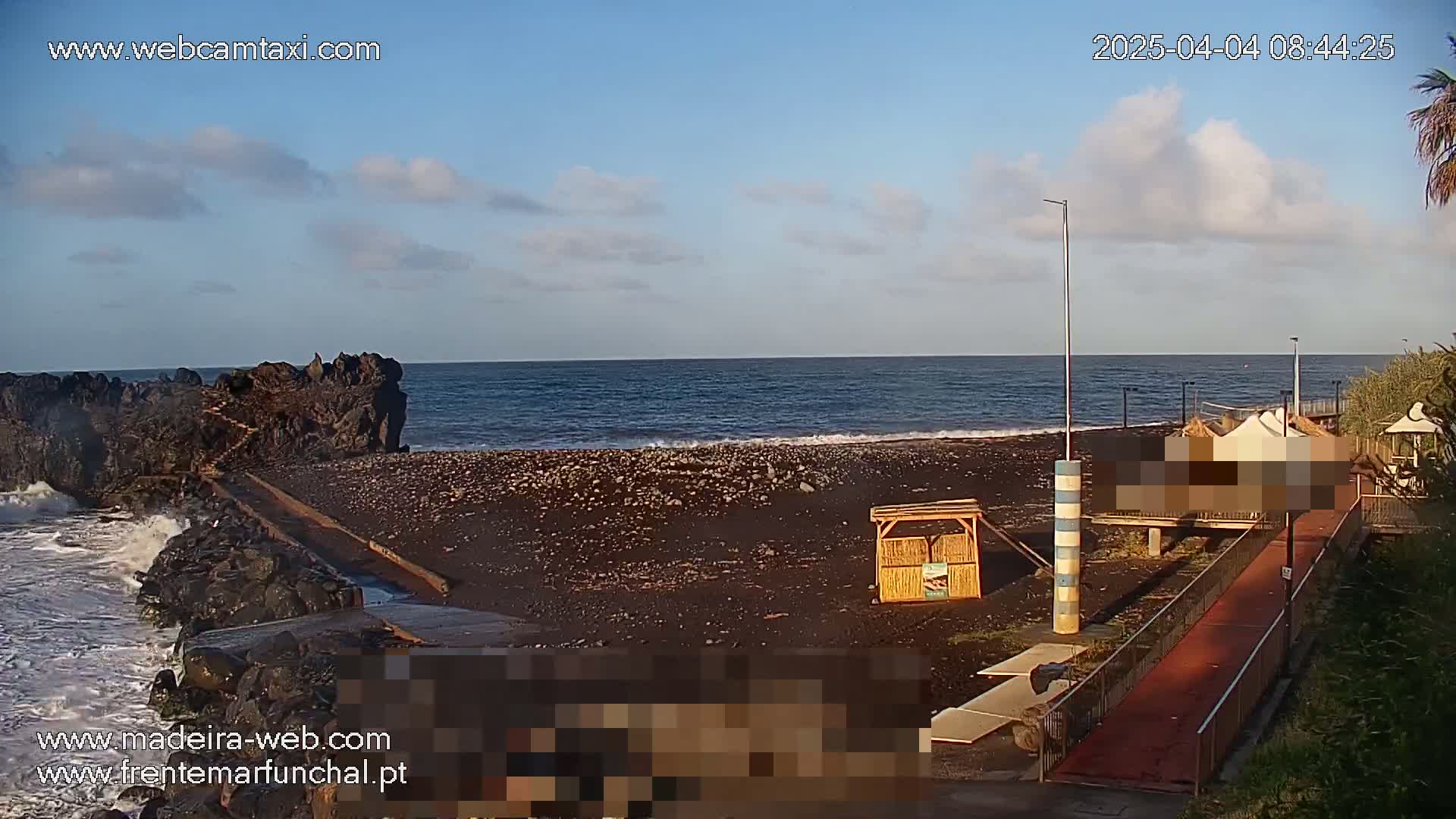 A dark-sand beach with a rocky outcrop at one end, under partly cloudy skies, with calm waves lapping at the shore.
