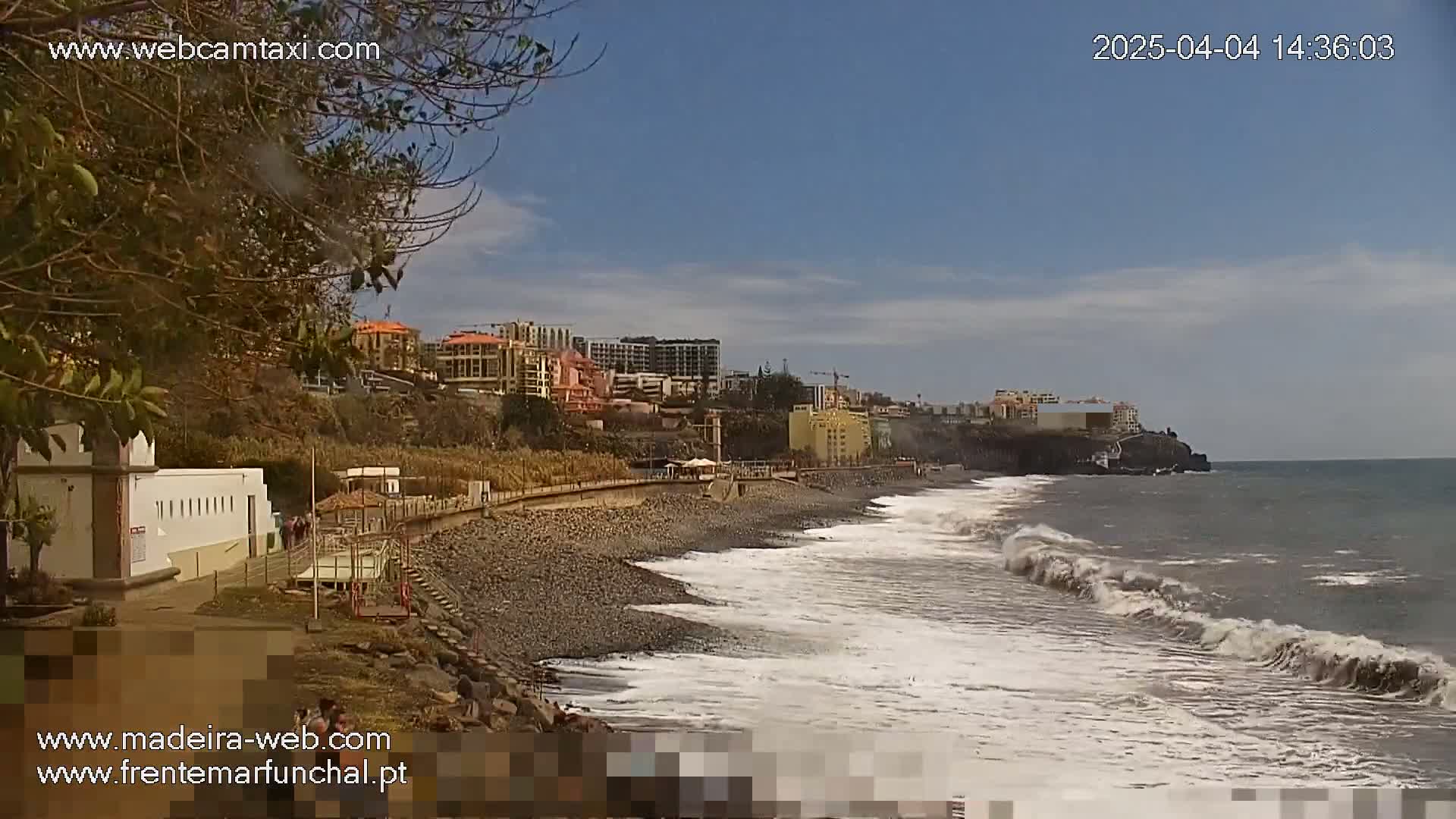A rocky beach with moderate waves laps against the shore near buildings on a partly cloudy day.