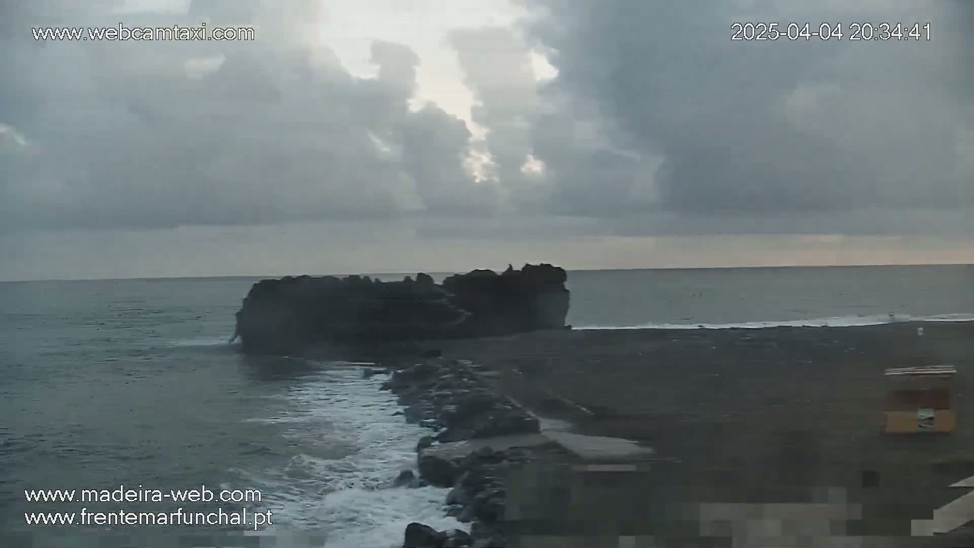 A dark, rocky outcrop sits in the ocean under a cloudy sky, with waves crashing against a nearby shore.