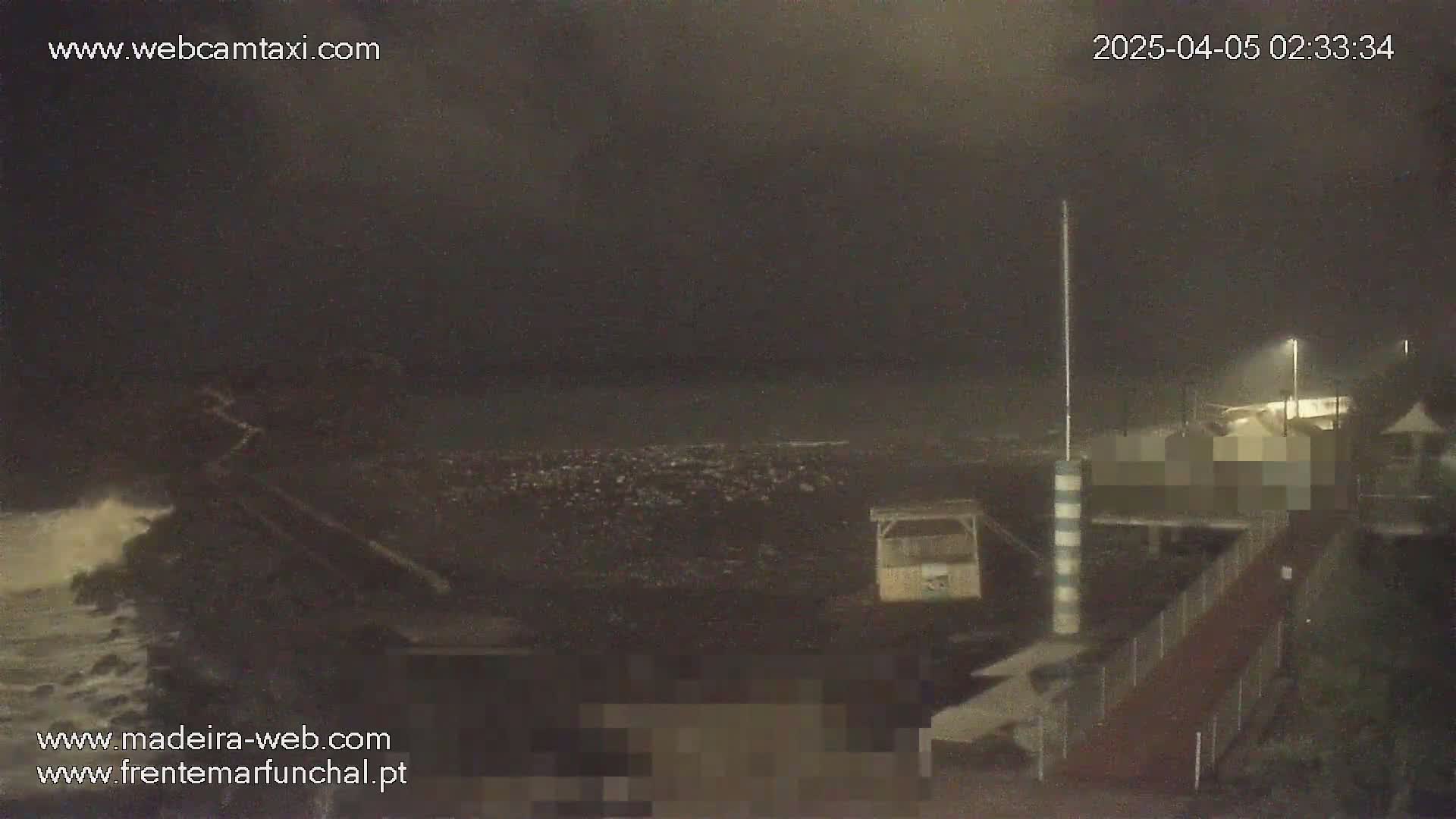 A nighttime, dark, and stormy ocean scene shows waves crashing against a rocky shoreline near a pier with structures, under a dark sky.