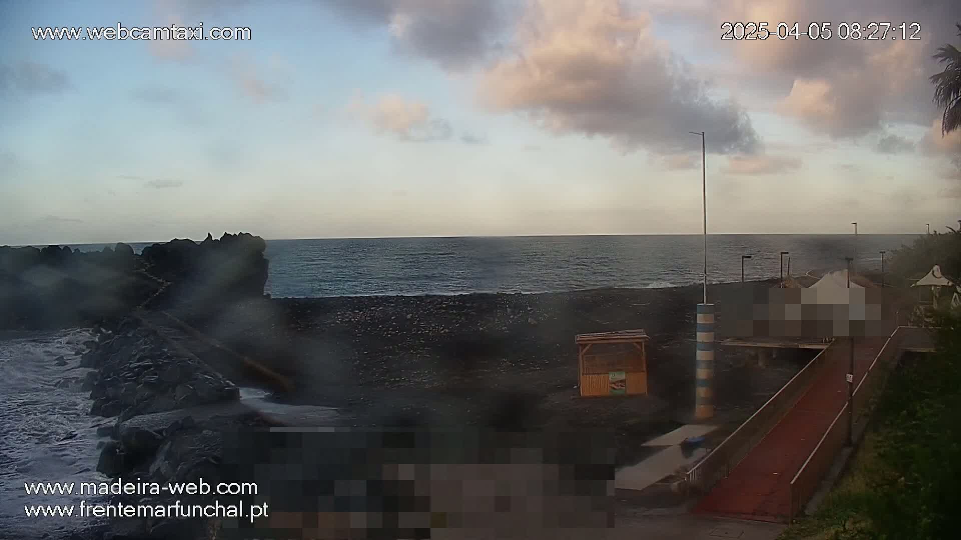 A partly cloudy sunrise over a dark-sand beach and calm ocean, viewed from a seaside walkway.
