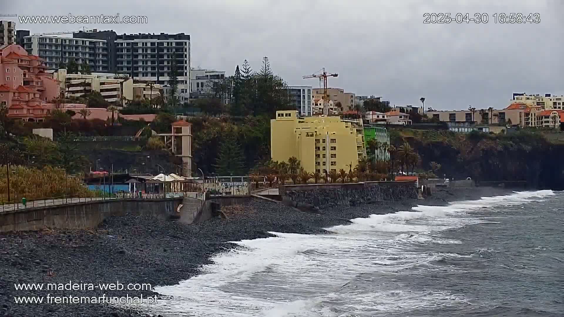 A black sand beach with moderate waves laps against a seawall separating it from buildings and a hillside under an overcast sky.