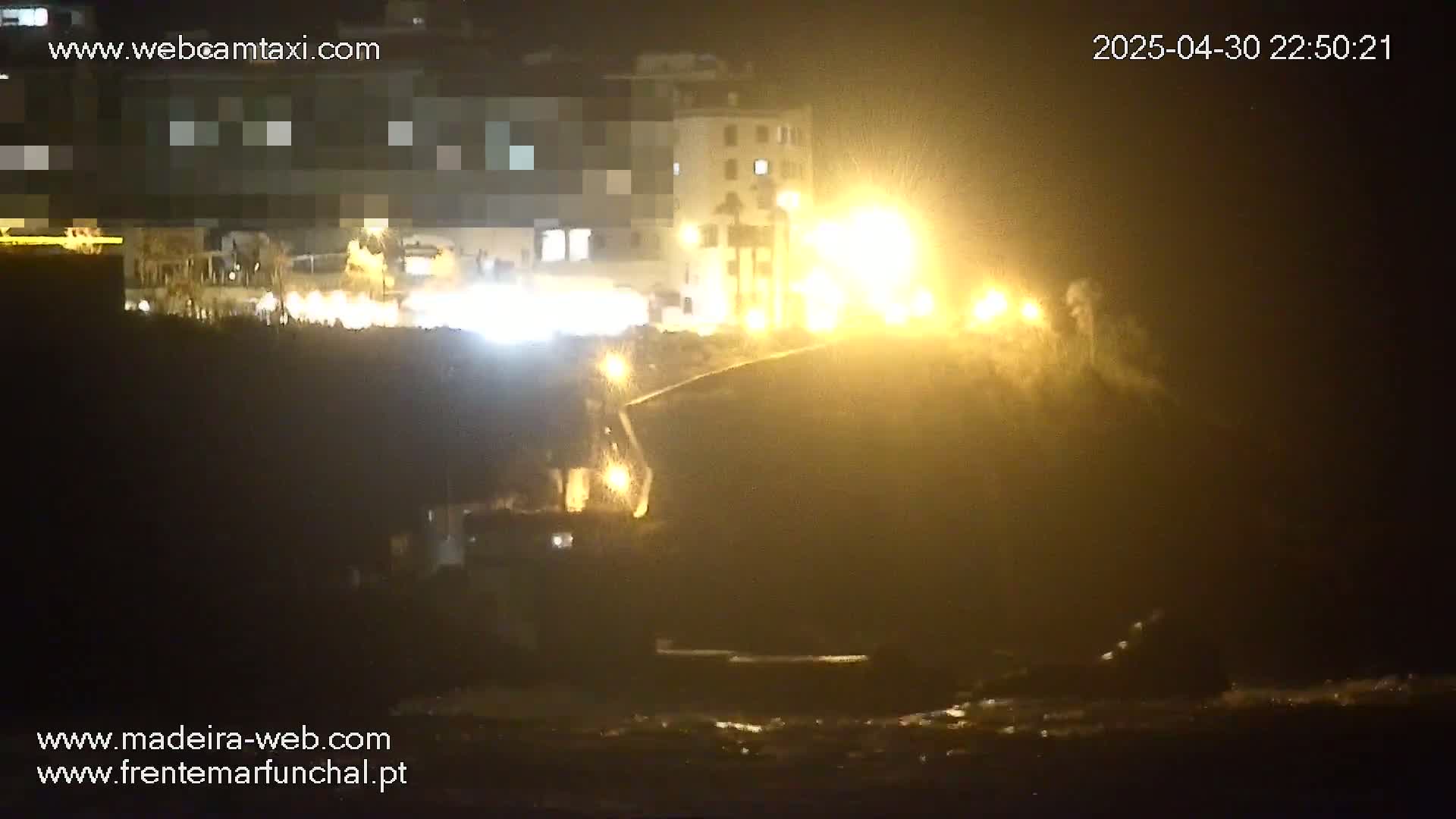 A nighttime, dimly lit scene shows a dark body of water near a shoreline with buildings and bright lights visible in the background.