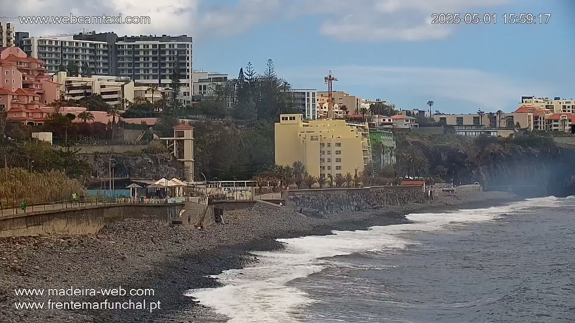 A rocky beach with gentle waves laps against the shore, adjacent to a boardwalk and several buildings under a partly cloudy sky.