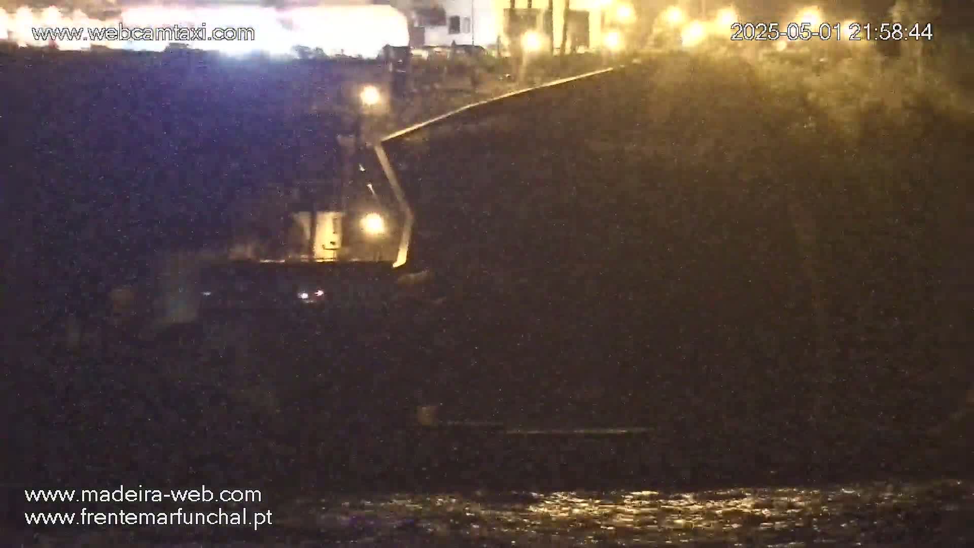A dark, nighttime scene shows a vehicle near a body of water under a light rain.