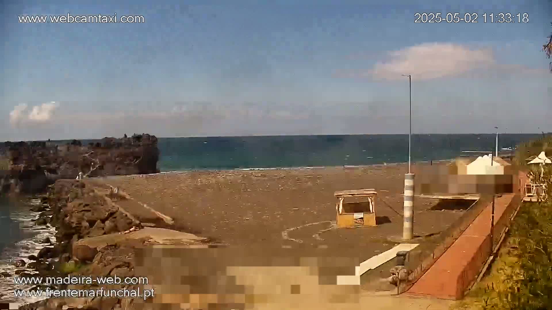 A mostly sunny day at a dark-sand beach shows a rocky point jutting into a calm ocean, with a few beachgoers visible and a boardwalk leading to a small structure.