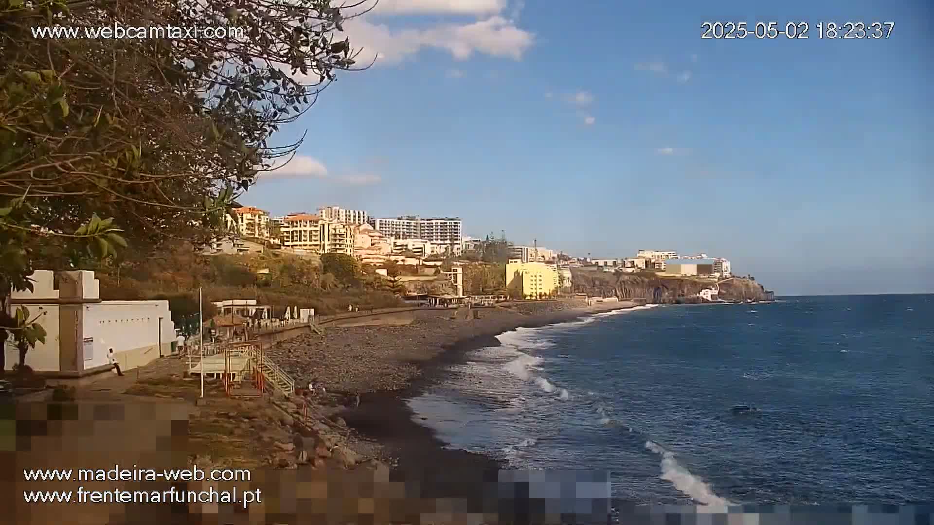 A dark-sand beach stretches along a coastline, with buildings and a cliff in the background under a partly cloudy sky.