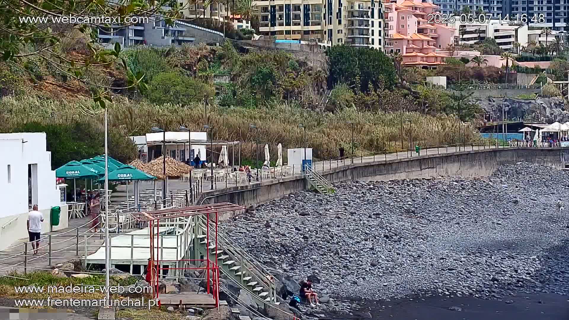 A rocky shoreline with a restaurant and walkway overlooking a dark-colored beach, under a sunny sky.