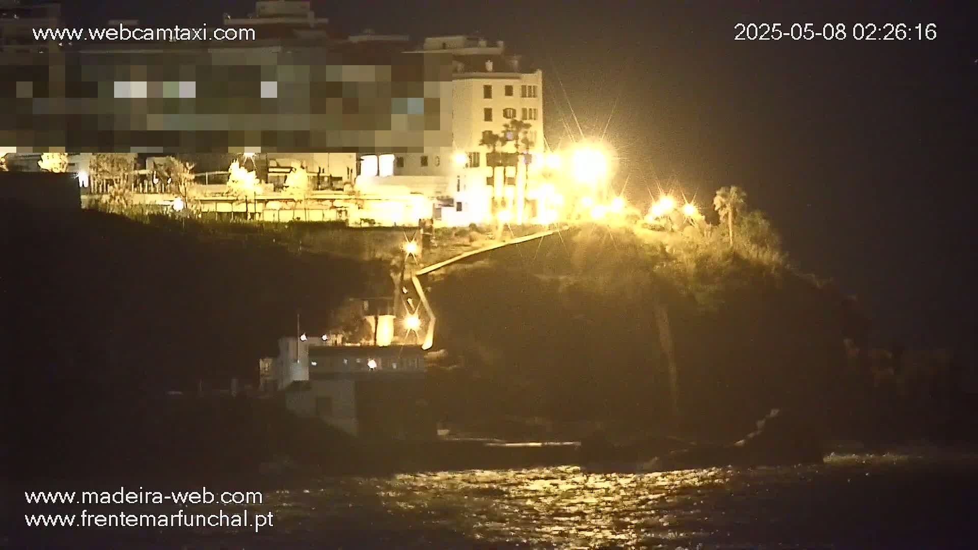 A nighttime coastal scene shows a building perched atop a cliff overlooking a dark, wavy ocean, illuminated by streetlights.